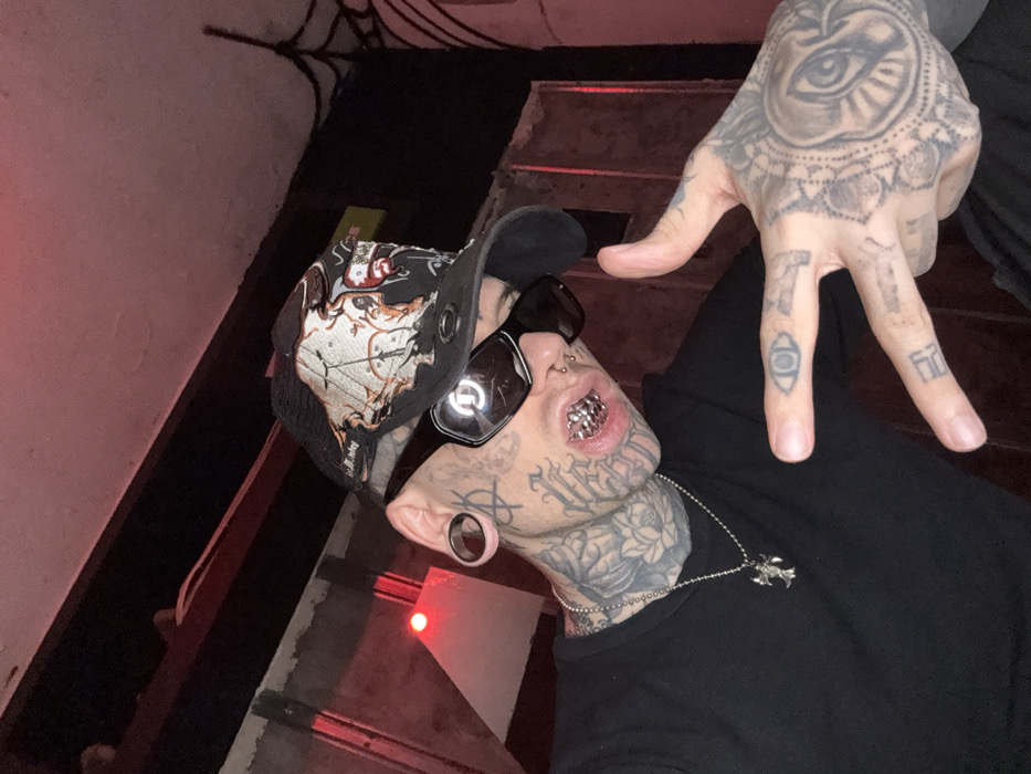 The image presents a close-up, angled portrait of a single individual within a dimly lit indoor setting. The subject, who appears to be male, is extensively modified with black and grey tattoos covering the face, neck, and hands, displaying intricate patterns and symbols including what resembles a lotus flower on the neck and an 'all-seeing eye' on the left hand. He wears dark, rectangular sunglasses, a black baseball cap with intricate graphics, including a skull motif, worn backward, and large circular ear gauges in both earlobes. Silver grillz are visible on both the upper and lower teeth as the mouth is slightly open. A silver ball-chain necklace with a winged, gothic-style pendant hangs around his neck. The individual's left hand (viewer's right) is raised, with the index and middle fingers extended in a two-finger gesture. The background is dark, featuring a red light source on the upper left, dark structural beams overhead, and a black spider web decoration in the upper right corner, indicative of a themed environment. The specific location within the city of Buenos Aires, Argentina, cannot be determined from the visual information presented.