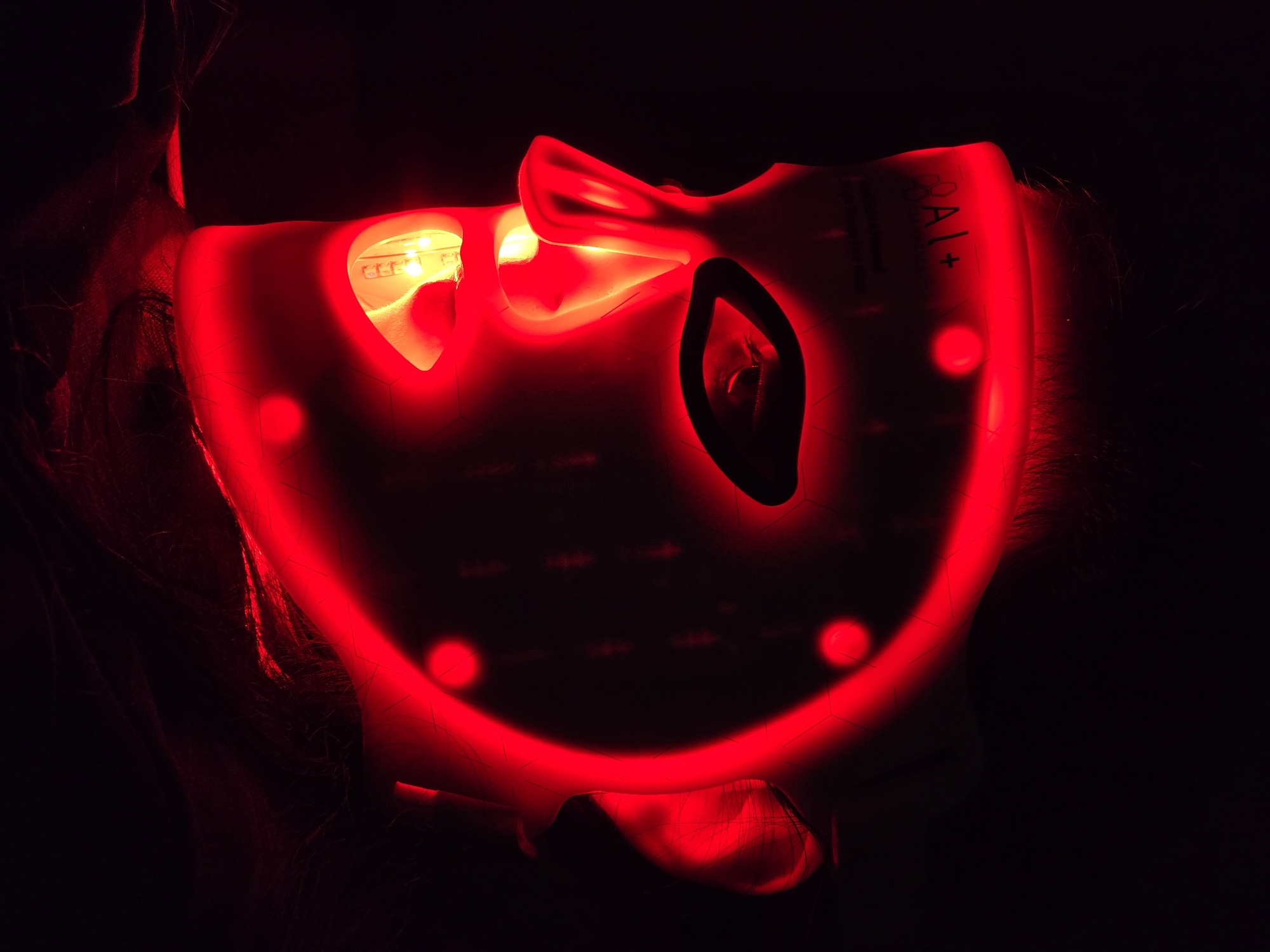 A close-up, dimly lit image shows a person's face obscured by a red light therapy mask. The mask is the primary light source, emitting a vivid red glow that illuminates parts of the wearer's skin and hair. The person's right eye is partially visible through an opening in the mask, while their hair, dark and flowing, is highlighted by the red light around the edges of the mask. The mask itself is white or light-colored, but entirely bathed in red light, revealing internal light-emitting diodes (LEDs) around the eyes, nose, and mouth openings. Several distinct circular red lights are visible embedded in the mask's surface, particularly along the chin and forehead areas. The background is completely black, emphasizing the glowing mask and the person's form within the darkness. On the upper right side of the mask, the partial text "OAI+" is visible. The scene suggests an indoor setting, likely at night or in a darkened room, focused on a beauty or wellness treatment.