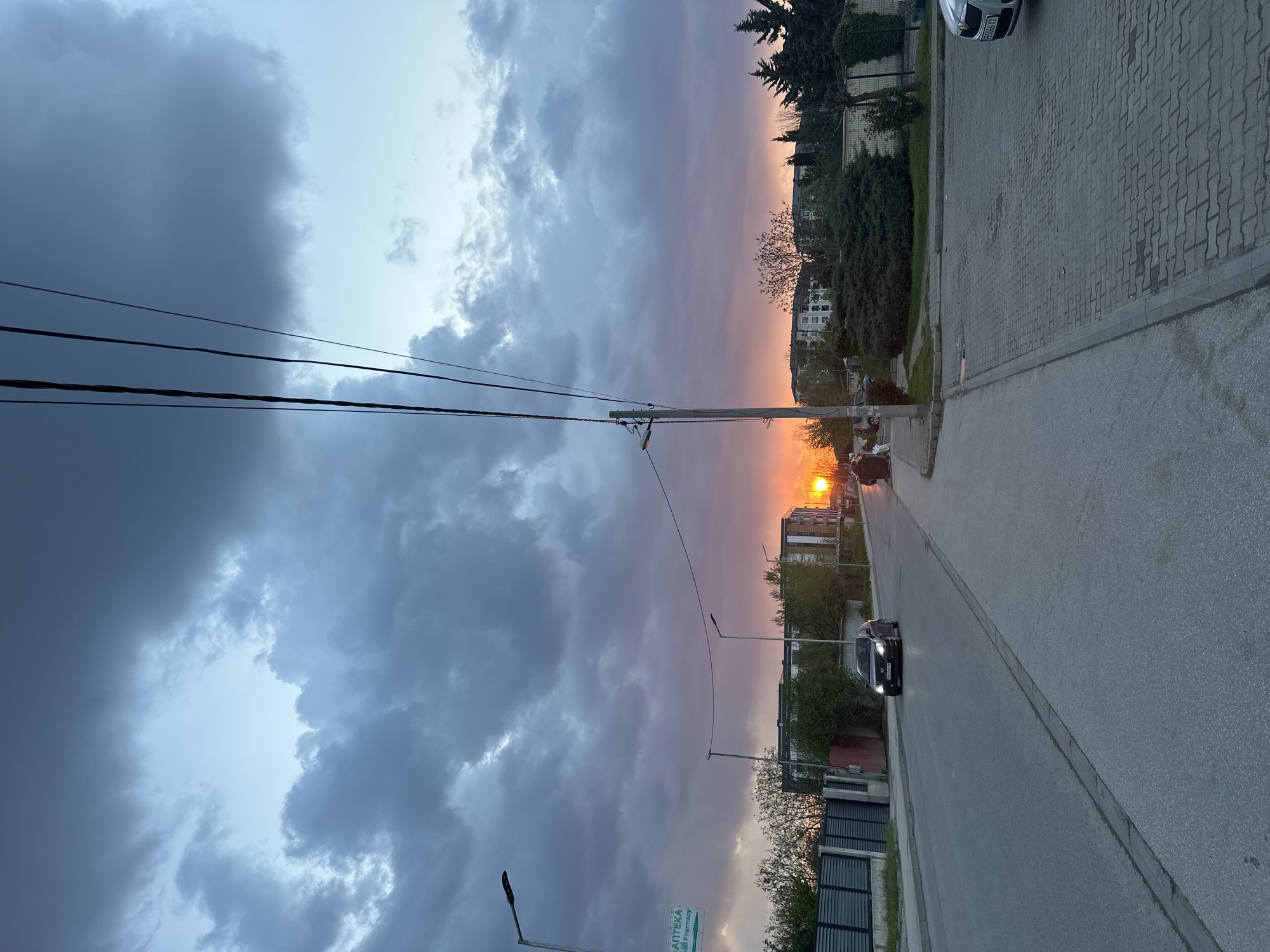 The image captures an urban landscape at sunset, likely in Plovdiv, Bulgaria. The sky is filled with dramatic clouds, ranging from dark storm clouds on the left to lighter, sunset-tinted clouds on the right. The sun is visible near the horizon, creating an orange and pink glow in the sky.
A street runs through the center of the image, with a car traveling along it. Buildings and trees line the sides of the street. Utility poles with wires stretch across the sky.
A sign is visible on the left side of the street, partially obscured, but with the word "APTEKA" (pharmacy in Bulgarian) visible. The street appears paved, and a paved area is also seen at the top of the image, adjacent to the trees and buildings.