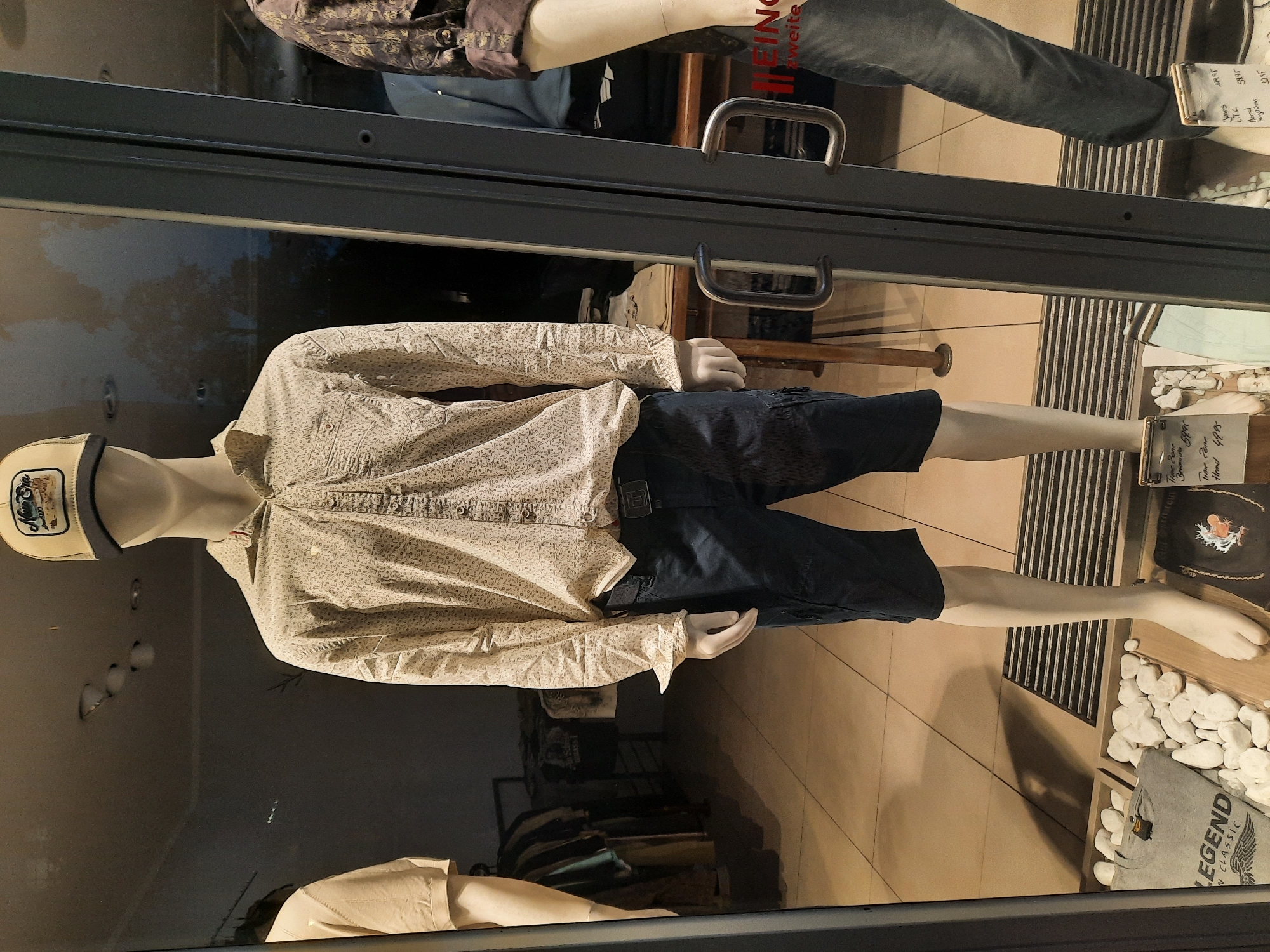 A male mannequin, positioned in the center of a store window in Rosenheim, Germany, is dressed in a patterned long-sleeved shirt, dark cargo shorts, a black belt with a silver buckle, and white gloves. The mannequin wears a tan baseball cap with a dark brim and a graphic logo. To the left, another mannequin, partially visible, wears a light-colored short-sleeved shirt. To the right, a third mannequin, also partially visible, wears a patterned long-sleeved shirt and jeans. In the foreground, near the base of the central mannequin, a gray t-shirt with the text "LEGEND CLASSIC" and a winged emblem is displayed amongst white stones. A black fabric item with an illustration is also visible. Two price tags are present; one displays "Time Zone Brownies 39.95" and "T-Shirt 49.95," and another, to the right, lists "Jeans (TC) 59.95" and "Head by Wear 27.95." The store window features two vertical metal handles.