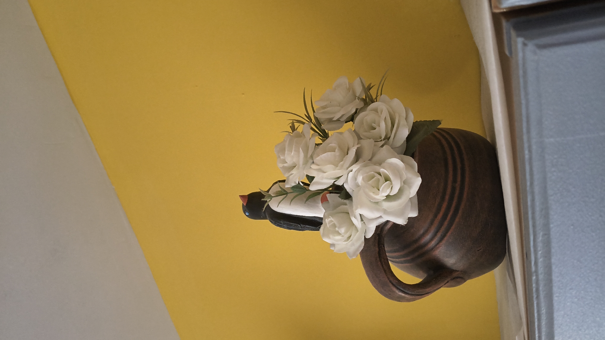 In Shumen, Bulgaria, on a surface likely a shelf or countertop, a brown clay jug with a handle holds a bouquet of artificial white roses. Perched atop the roses is a small, decorative figurine of a penguin with black and white plumage, a red beak, and orange feet. The background is a bright yellow wall meeting a light-colored ceiling at an angle. The lower portion of the image shows the edge of a gray surface, possibly furniture.