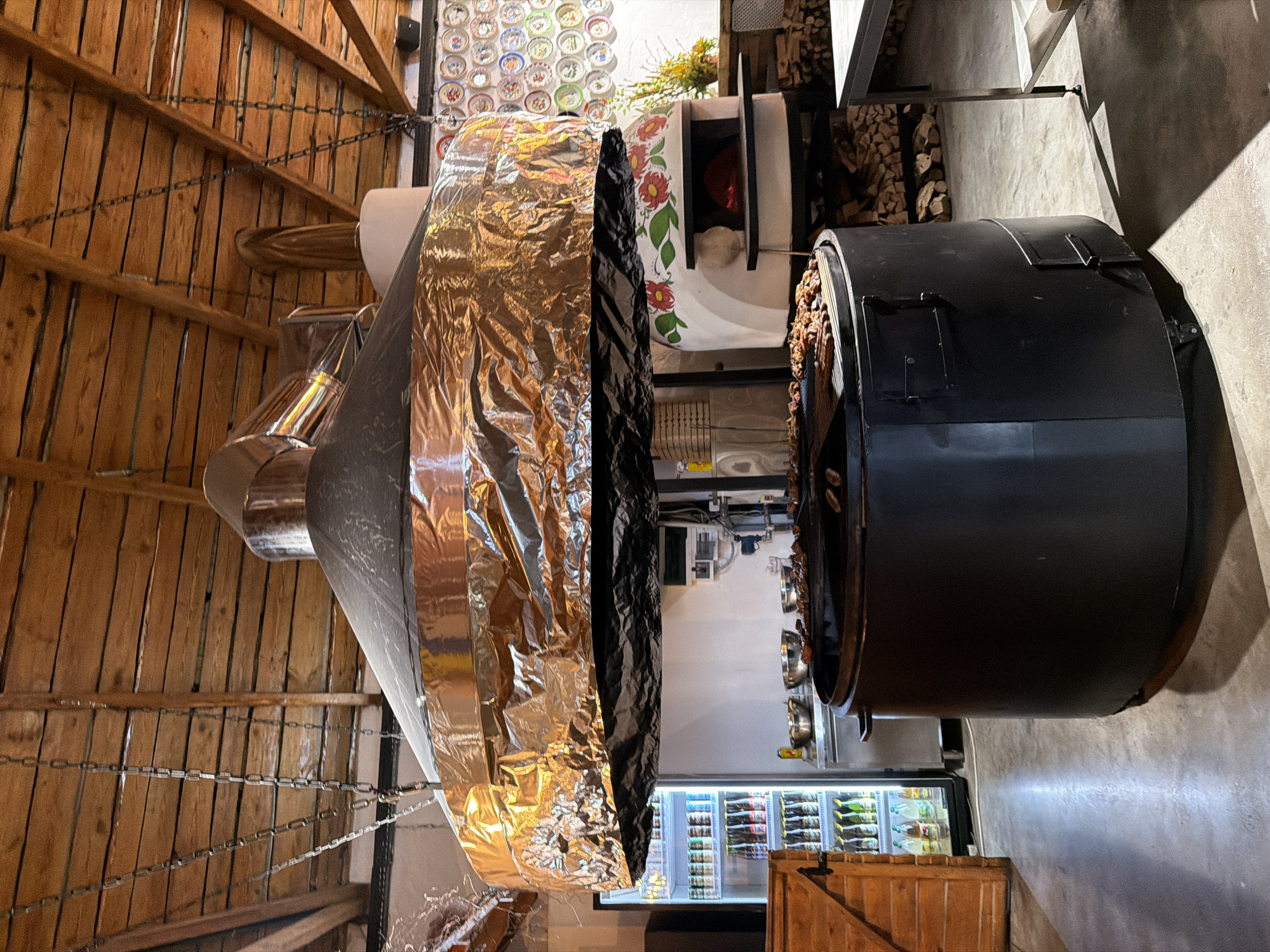 In Velykyi Khodachkiv, Ukraine, an indoor scene depicts a large, circular, black barbecue pit situated centrally. Above the pit hangs a substantial ventilation hood, partially covered in reflective foil. The pit contains numerous pieces of meat cooking on a grill grate. To the left of the pit, a glass-doored refrigerator is filled with bottles. Behind the barbecue, a white oven, adorned with painted red and green floral designs, is visible with a glowing red interior. To the right of the oven, stacks of firewood are piled. The ceiling is constructed of wooden planks supported by beams, with chains hanging down. The floor is concrete. The scene appears to be a commercial kitchen or dining area.