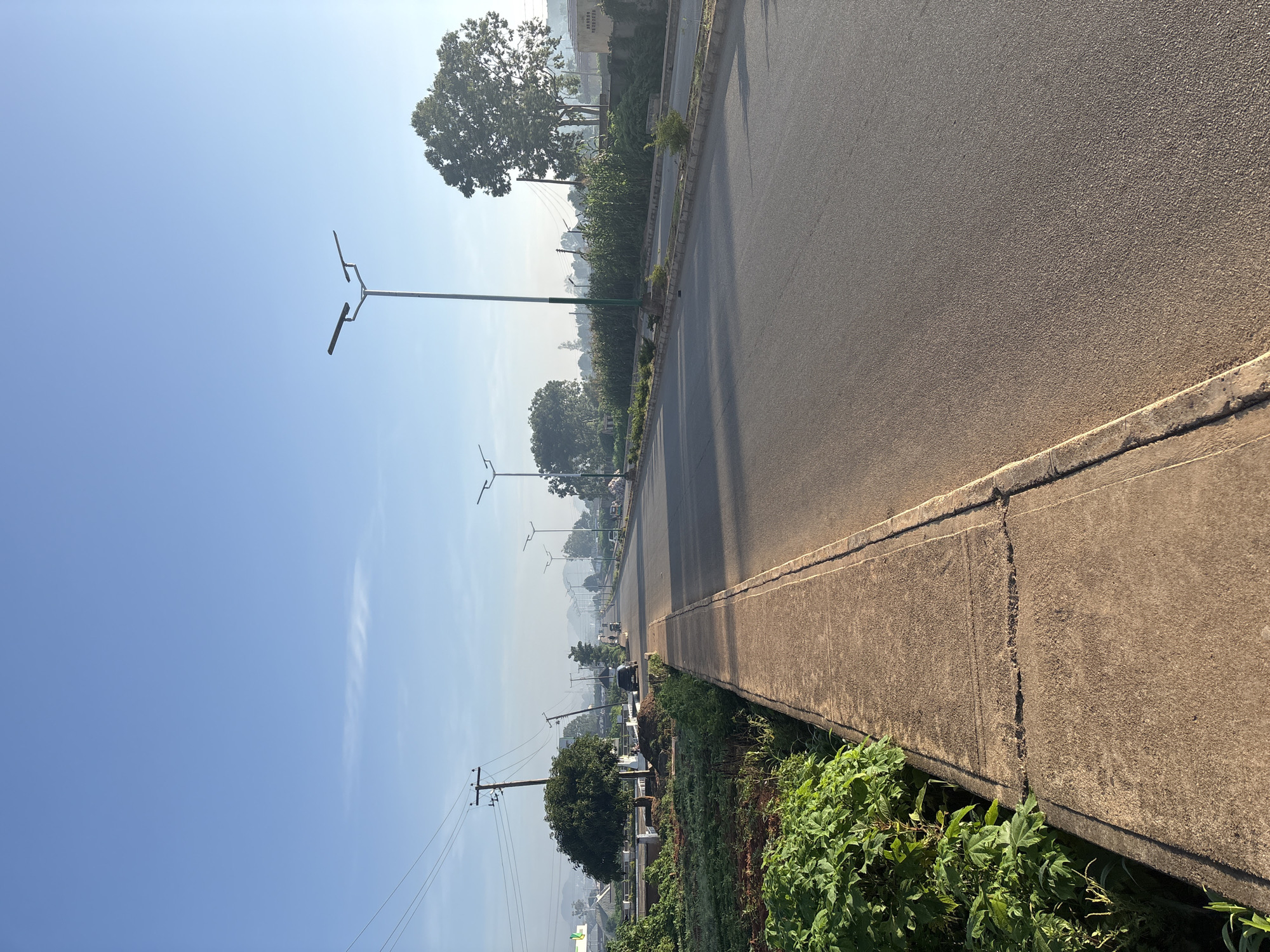 In Bukuru, Nigeria, on a clear morning, a paved road stretches towards a distant horizon under a bright blue sky. The road is bordered by a concrete sidewalk on the left, which is overgrown with lush green foliage in some areas. Along the sidewalk, there are several trees and a few streetlights, spaced evenly apart, illuminating the path. On the right side of the road, there are more trees, some of which are quite large and mature. The road is mostly empty, with a few vehicles visible in the distance, suggesting a quiet start to the day. The scene is tranquil and serene, with the natural beauty of the vegetation contrasting with the man-made road and streetlights.
