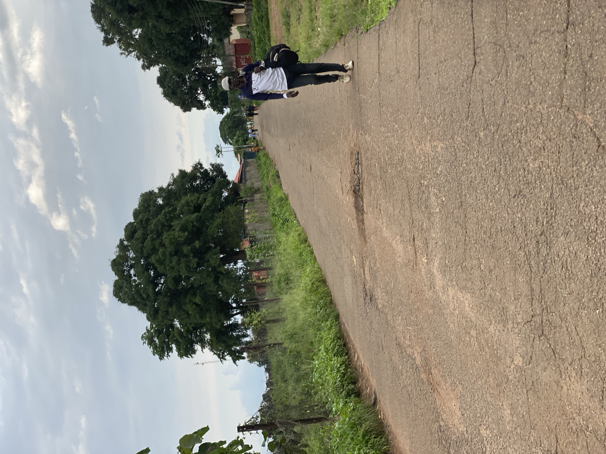 A man walks down a cracked asphalt road in Maigizo, Nigeria, carrying a black bag over his right shoulder. He is wearing a white, long-sleeved shirt with blue accents and the number "2" on the left sleeve, dark pants, and light-colored sneakers. A beige baseball cap covers his head. The road is lined with dense green grass and foliage on the left, separated by a wire fence and wooden posts. On the right, the grass continues to the edge of the road, with trees and buildings visible in the distance. The sky is overcast with scattered white clouds. Several other people are visible further down the road, walking in the same direction as the main subject.