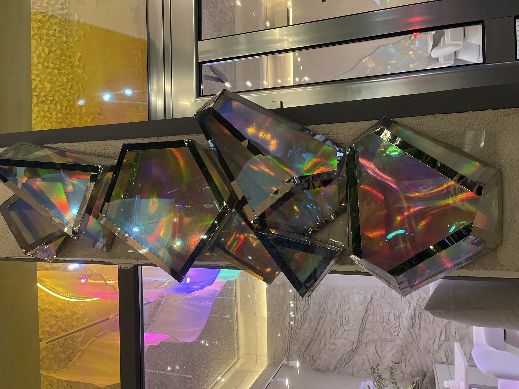 The image shows a decorative art installation. It is comprised of several geometric, mirrored shapes mounted on a textured, off-white wall. The mirrored surfaces reflect surrounding light and colors, creating an iridescent effect. Adjacent to the wall with the mirrored installation are two windows with reflective glass; through these windows, interior elements can be seen, including a yellow-toned wall and a structure illuminated with a pink-purple light. The presence of furniture partially visible at the bottom right suggests an indoor space. The location is Krasnodar, Russia, according to the task.