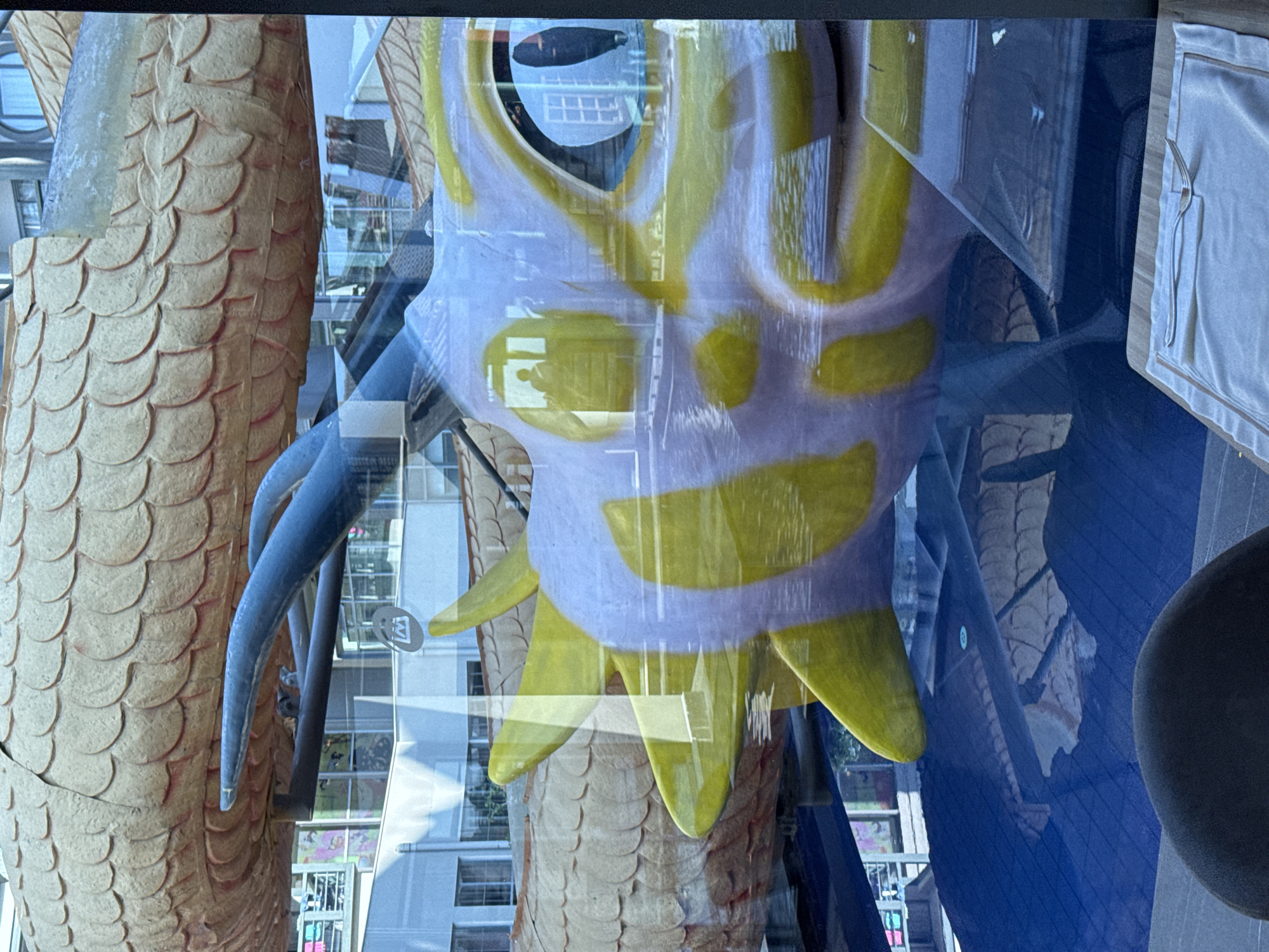 The image is a close-up shot of a large, stylized dragon float with yellow and white markings. The dragon's large eye is visible, reflecting a building with windows and a dark figure. The float appears to be on or near water, which is a deep blue and reflects the underside of the dragon. In the foreground, a dark, rounded object, possibly a chair, is partially visible, along with a placemat and a fork. The background shows a building with multiple windows and a sign with a stylized "M". The scene is likely an outdoor setting, possibly a city park or event, but the location in Aksu, Turkey cannot be confirmed from the image alone.