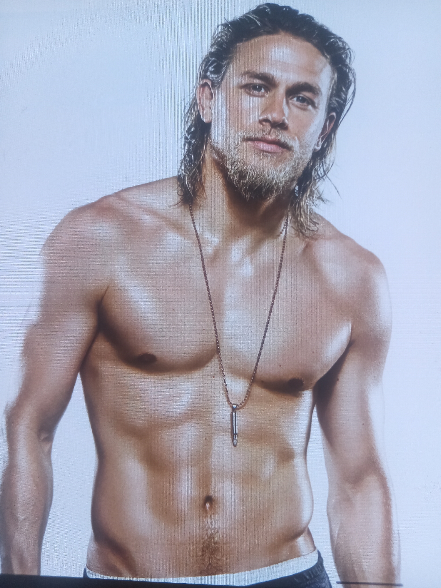 This media file is a portrait of a man, likely an actor, posed against a plain white background. The subject is shirtless, revealing a muscular physique. He has medium-length, dark, wavy hair that is styled away from his face, and a light beard. A silver-colored pendant, shaped like a bullet casing, hangs from a thin chain around his neck. The lighting highlights the contours of his chest, abdomen, and arms, suggesting a studio setting. The man's expression is neutral, with his gaze directed towards the viewer. The overall aesthetic is one of deliberate presentation, emphasizing the subject's physical form and possibly his character. There is no discernible text in the image.