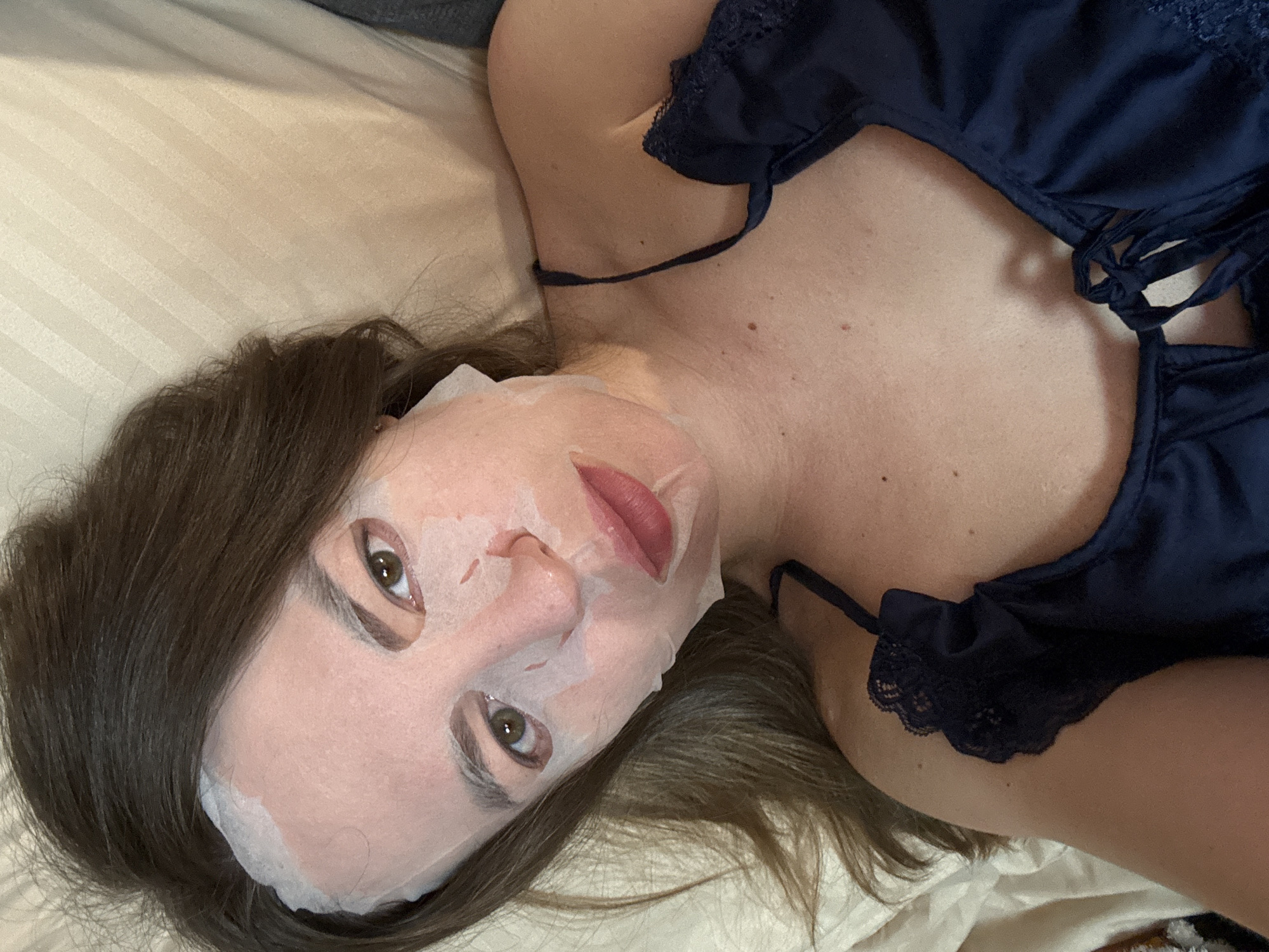 A woman lies on a cream-colored bed wearing a blue nightgown with lace trim. A white sheet mask covers her face, except for her eyes and lips. She has brown hair. No information about the location is discernible from the image.