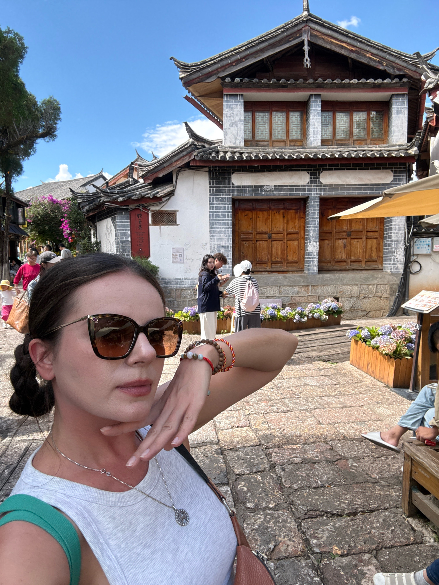 A woman, facing the camera and taking a selfie, stands on a cobblestone street in Gucheng District, China. She has her right hand near her face, with her fingers delicately touching her chin. She is wearing large, brown-rimmed sunglasses and a light-colored tank top with a green strap. A silver necklace with a circular pendant is visible around her neck. Behind her, traditional Chinese architecture with dark tiled roofs and wooden doors is prominent. Several people are in the background, including a group near the building and a child walking away from the camera on the left. Flower boxes filled with colorful blooms are placed along the street. The sky is blue with scattered clouds, indicating a bright, sunny day.