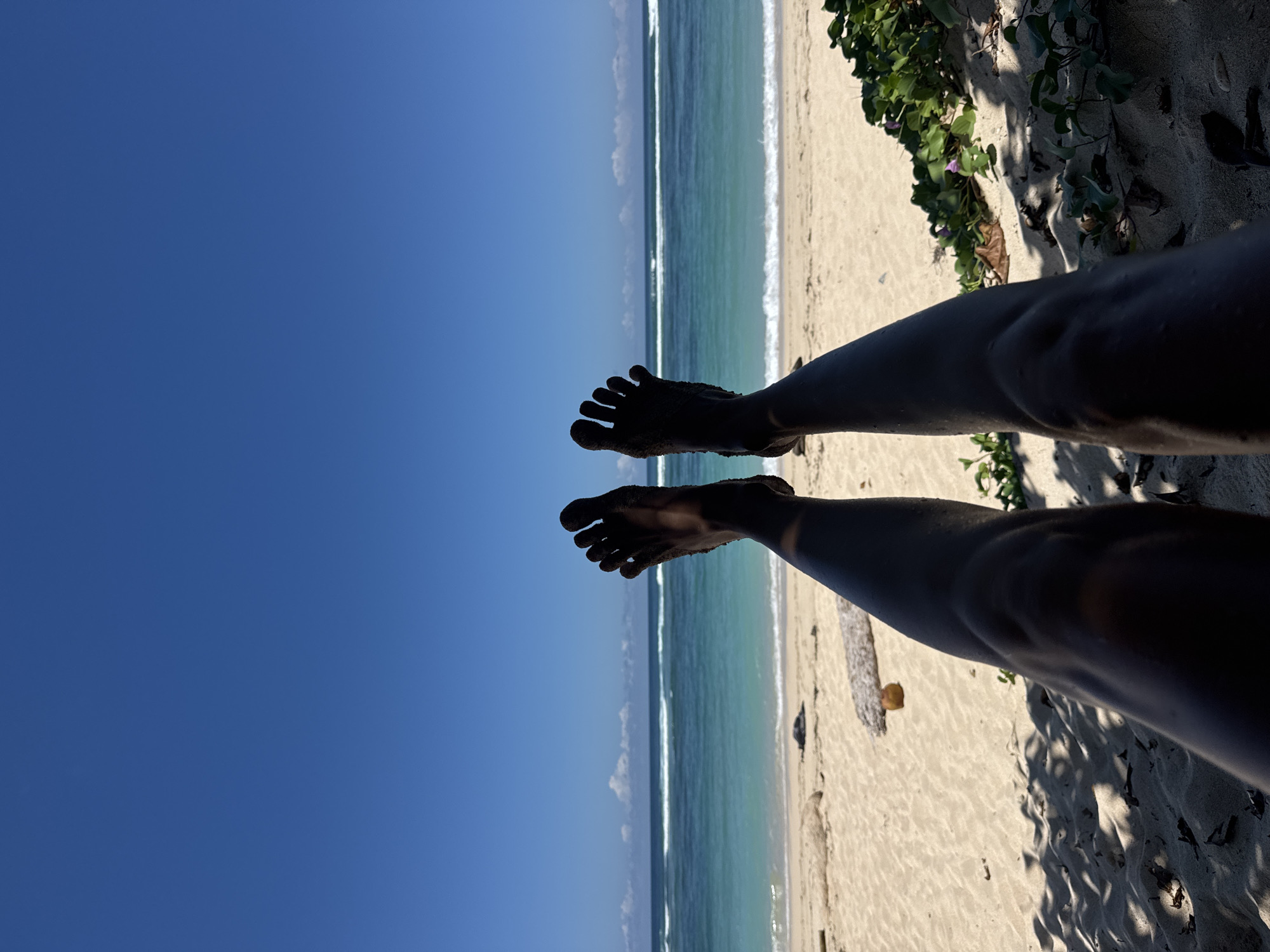 This media file captures a serene, first-person perspective of someone relaxing on a tropical beach in Kuta Selatan, Indonesia. The image is composed with the viewer's dark-skinned legs and sand-covered feet prominently in the foreground, silhouetted against the bright background. The feet appear relaxed, suggesting the person is lying down or reclining on the sand.
Beyond the feet, the scene unfolds into a vibrant seascape. To the left, a clear, deep blue sky dominates, while to the right, the turquoise water of the ocean meets a light-colored sandy beach. Gentle white waves are visible breaking near the shore, creating a distinct line where the water laps the sand.
Along the upper right and bottom right edges of the frame, green beach foliage, possibly low-lying vines, grows on the sand. These plants cast intricate shadows, indicating strong, overhead sunlight, suggesting the time of day is midday or early afternoon. A small, light-colored object, possibly a piece of driftwood or a fallen fruit, is visible on the sand near the right leg.
The scene embodies a sense of relaxation and vacation in a beautiful tropical setting, characterized by the clear sky, pristine sand, and the distinct colors of the ocean. There is no visible text in the image.