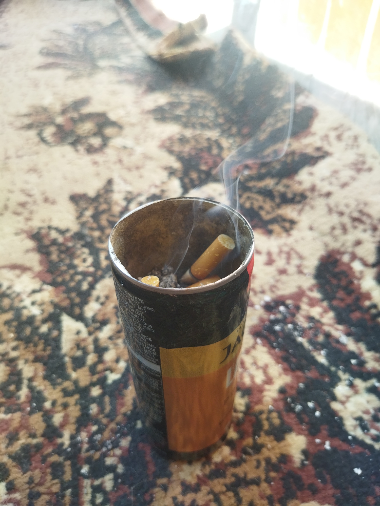 The image shows a makeshift ashtray, appearing to be a repurposed can, containing cigarette butts and emitting smoke. The can has labeling in a foreign language, including the text "JA". The ashtray is placed on a patterned rug or carpet with a blend of brown, tan, and black hues. The foreground is in focus, while the background is blurred, suggesting a shallow depth of field. The scene appears to be indoors. There is no information in the image about the city of Kramatorsk, Ukraine, or the presence of people.