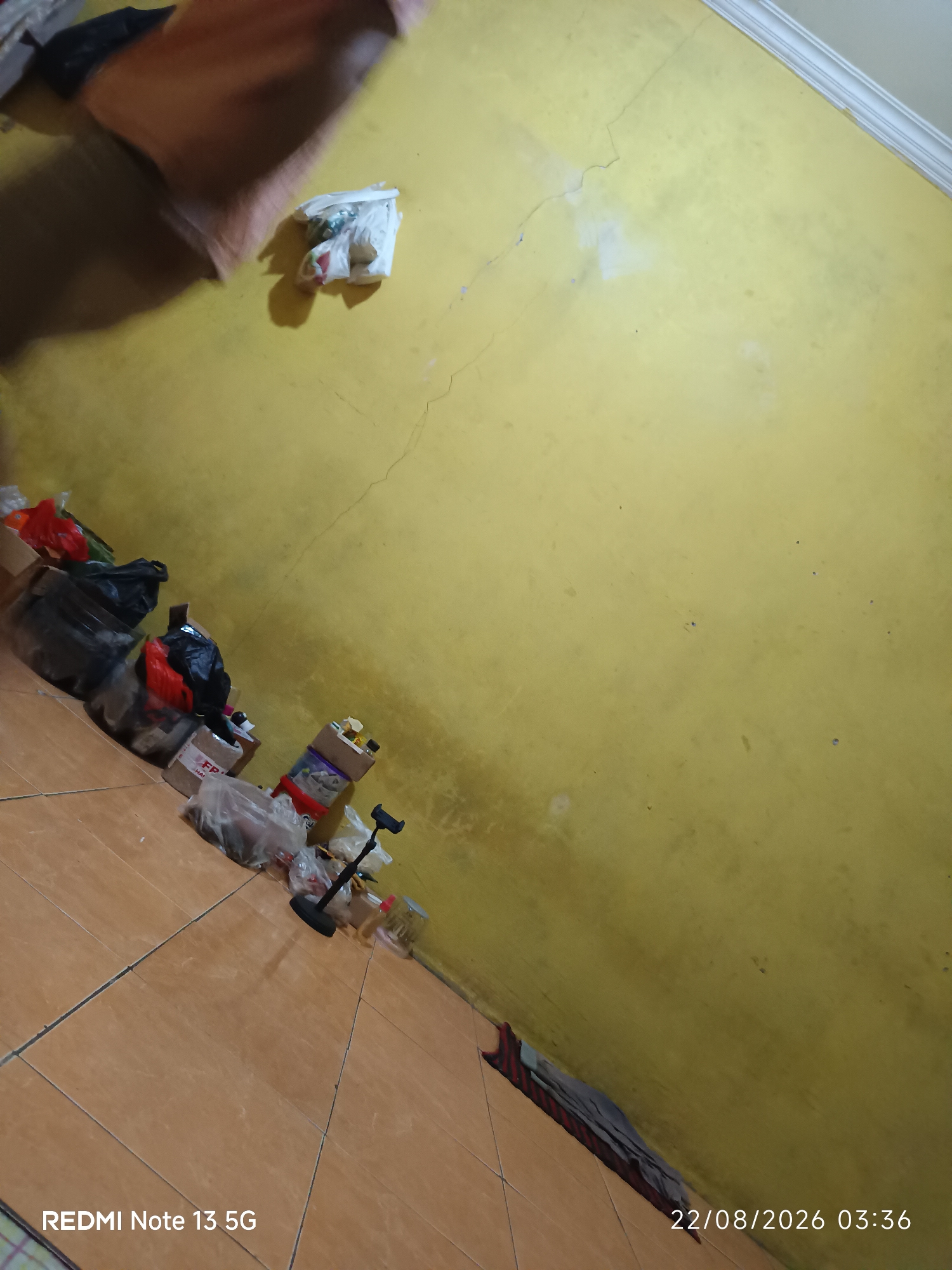 The image depicts a cluttered corner of a room with yellow walls and tiled flooring. In the foreground, on the left side, there is a collection of plastic bags and boxes piled on the floor. A black phone stand is visible near the center of the image. The wall in the background has some discoloration and cracks. A piece of orange fabric is draped over something at the top left of the image, casting a shadow. The bottom of the image has text overlays indicating "REDMI Note 13 5G" and a date and time stamp "22/08/2026 03:36". The scene appears to be indoors, possibly a storage area or a room with items left on the floor. The location is identified as Talaga Sari, Indonesia.
