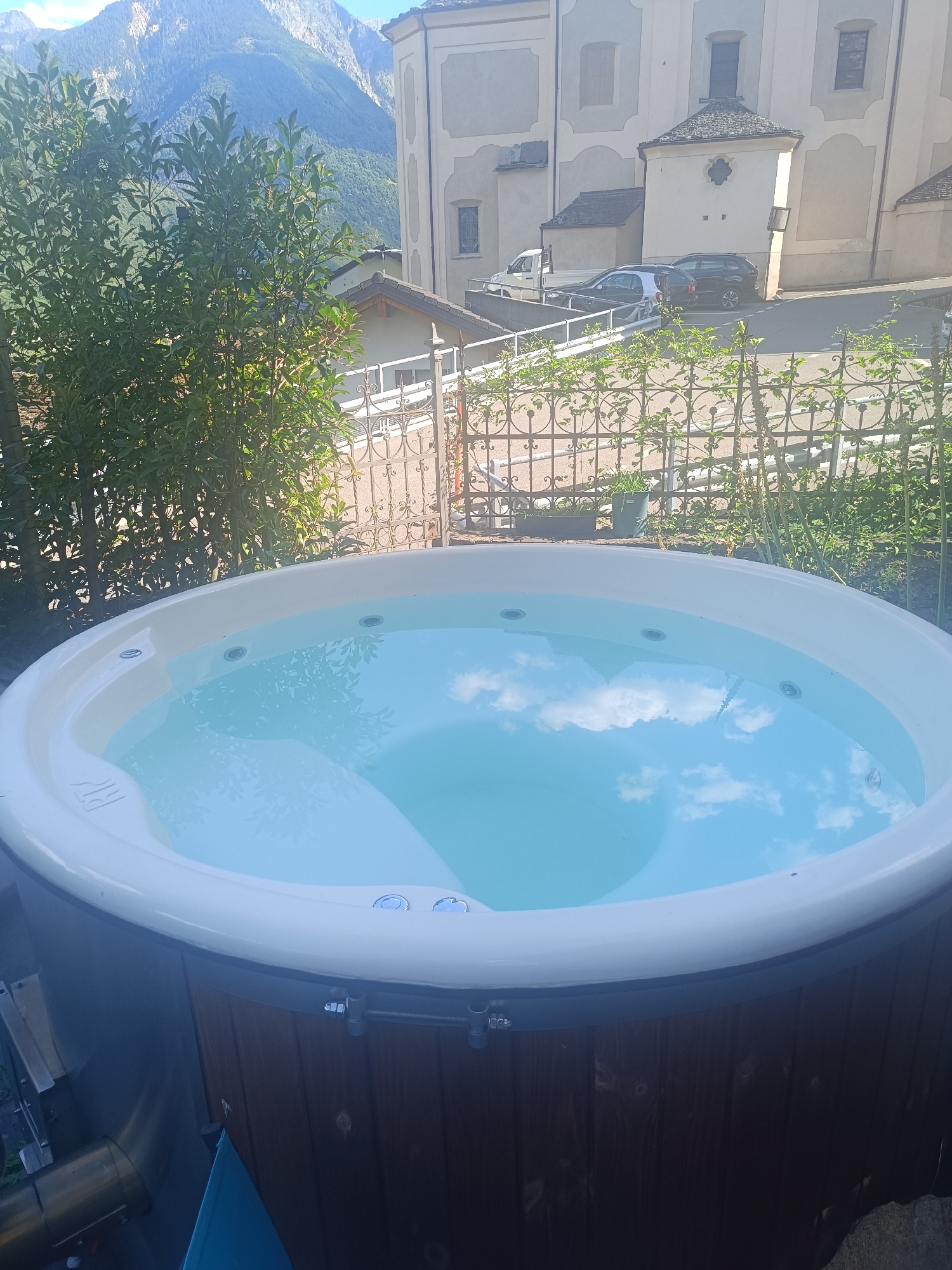 The image shows an outdoor hot tub with clear blue water, reflecting a bright sky with scattered clouds. The hot tub is round with a white rim and a wooden exterior. In the background, there is a white-gated fence and lush green trees. Beyond the fence, a building with an architectural design typical of alpine regions is visible, with a sloping roof and stone accents. The building appears to be a church or historical structure. Further in the distance, imposing mountains with rocky peaks and forested slopes rise under a clear blue sky. Several cars are parked on a paved area in front of the building. The scene suggests a peaceful setting in a mountainous region. The location is identified as Serravalle, Switzerland.