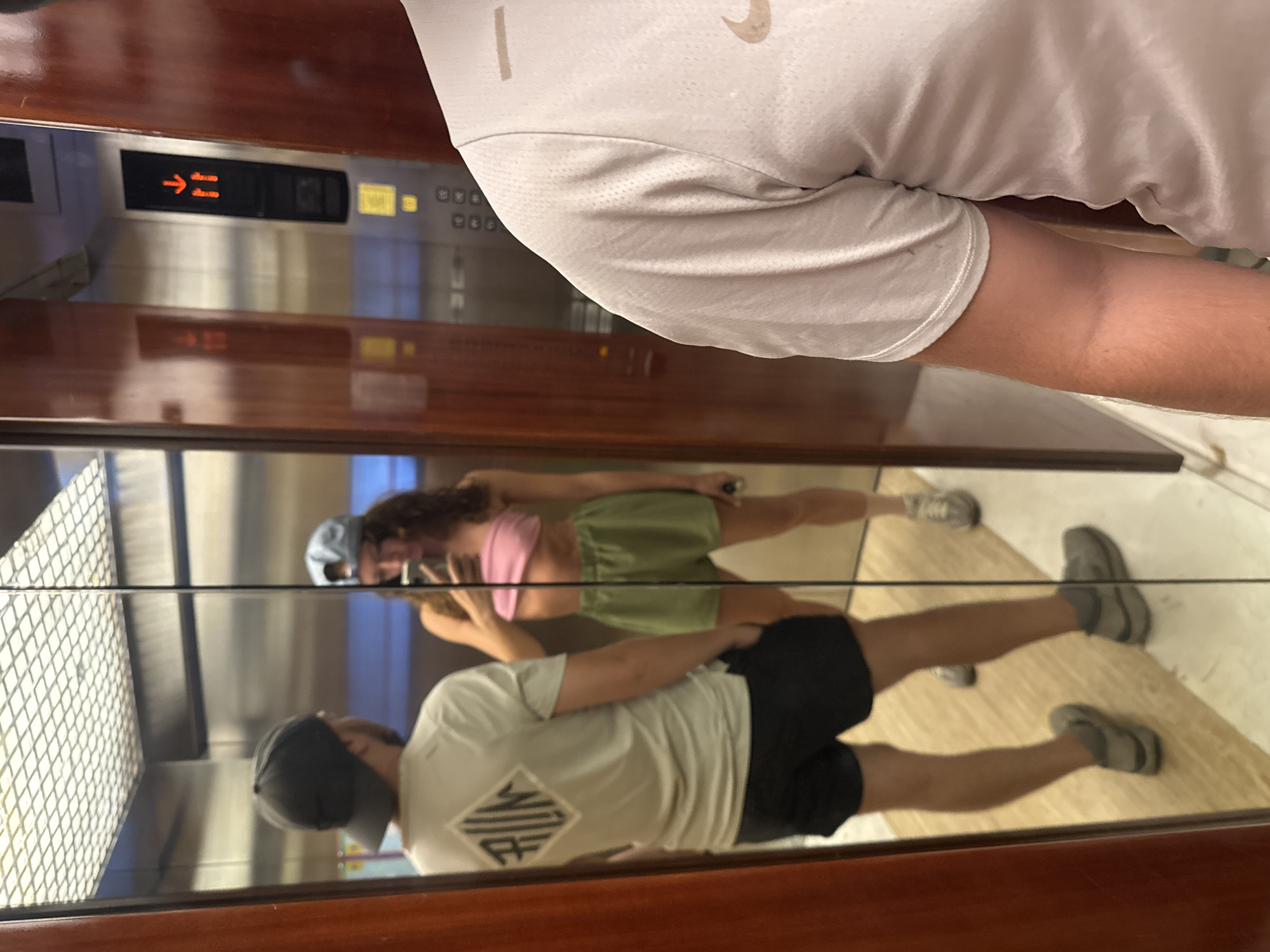 The image shows a reflection in a mirrored elevator in Đà Nẵng, Vietnam. Two people are visible in the reflection: a woman in a pink top and green shorts, and a man in a gray t-shirt, black shorts, and a cap. The elevator's floor display with an arrow is visible. The elevator's interior has metal walls and a panel of buttons.