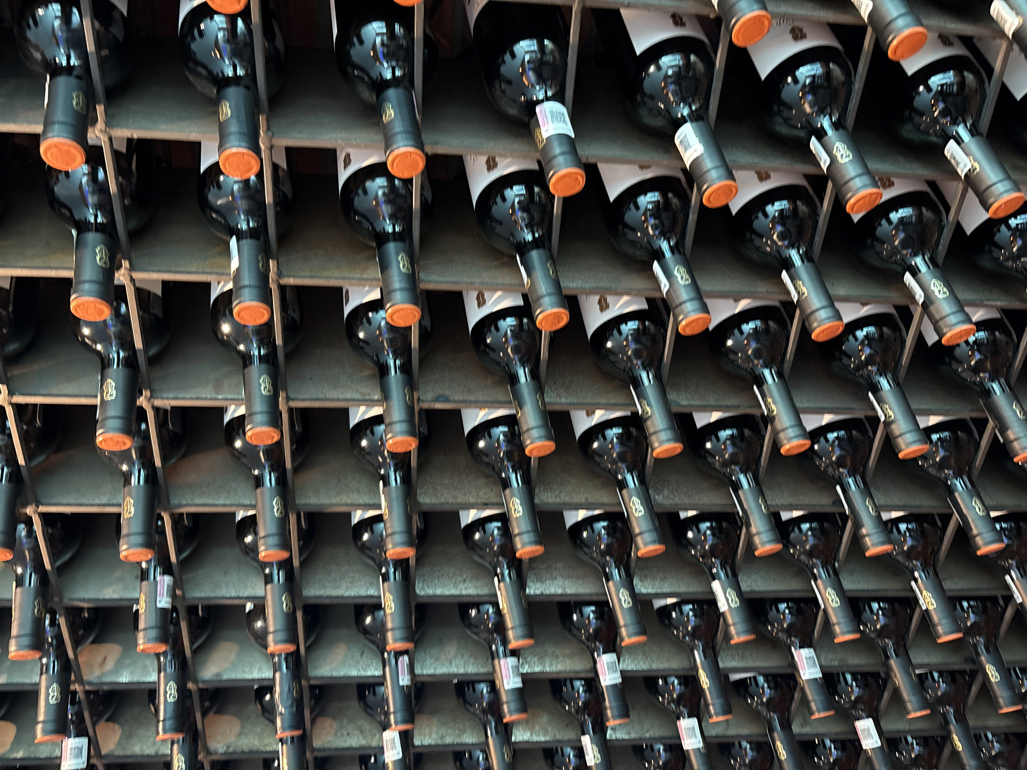A wine rack in Huixquilucan de Degollado, Mexico, holds numerous bottles of dark-colored wine horizontally in a metal grid. The bottles have orange-colored closures and white labels with possible branding.