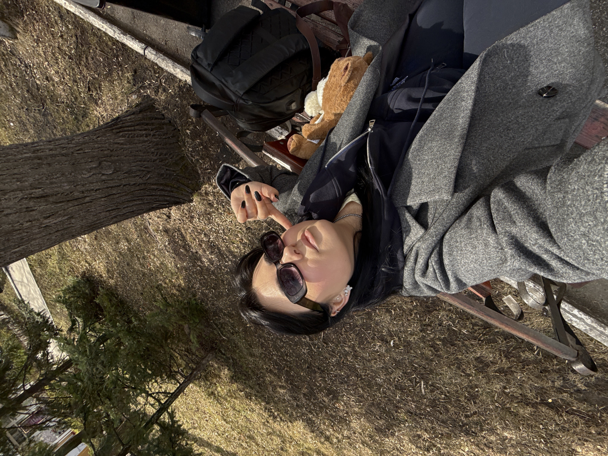 The image captures a diagonal, overhead view of a person relaxing on a wooden park bench in what appears to be an outdoor setting, possibly in Perm, Russia. The ground is covered with dried, light brown grass and foliage, suggesting a season like late autumn or early spring.
The primary subject is a young individual, likely female, with dark hair. She is dressed in a grey coat layered over a dark hoodie or jacket. Her hands, adorned with dark nail polish, are resting near her face. Dark sunglasses are pushed up onto her forehead, and small white, floral-shaped earrings are visible. Her face is angled slightly upwards, seemingly enjoying the bright sunshine.
Beside her on the bench are a black backpack and a small brown teddy bear, adding a personal touch to the scene. The bench itself features wooden slats and dark metal framing. To the left of the bench, a thick tree trunk with rough bark is prominently visible, rooted in the dry ground. Further in the background, other trees and shrubs contribute to the natural, park-like environment.
The strong sunlight creates soft shadows on the ground and highlights the subject, indicating a clear, sunny day, likely mid-morning or afternoon. The overall mood conveyed is one of relaxation and calm. No visible text is present in the image.