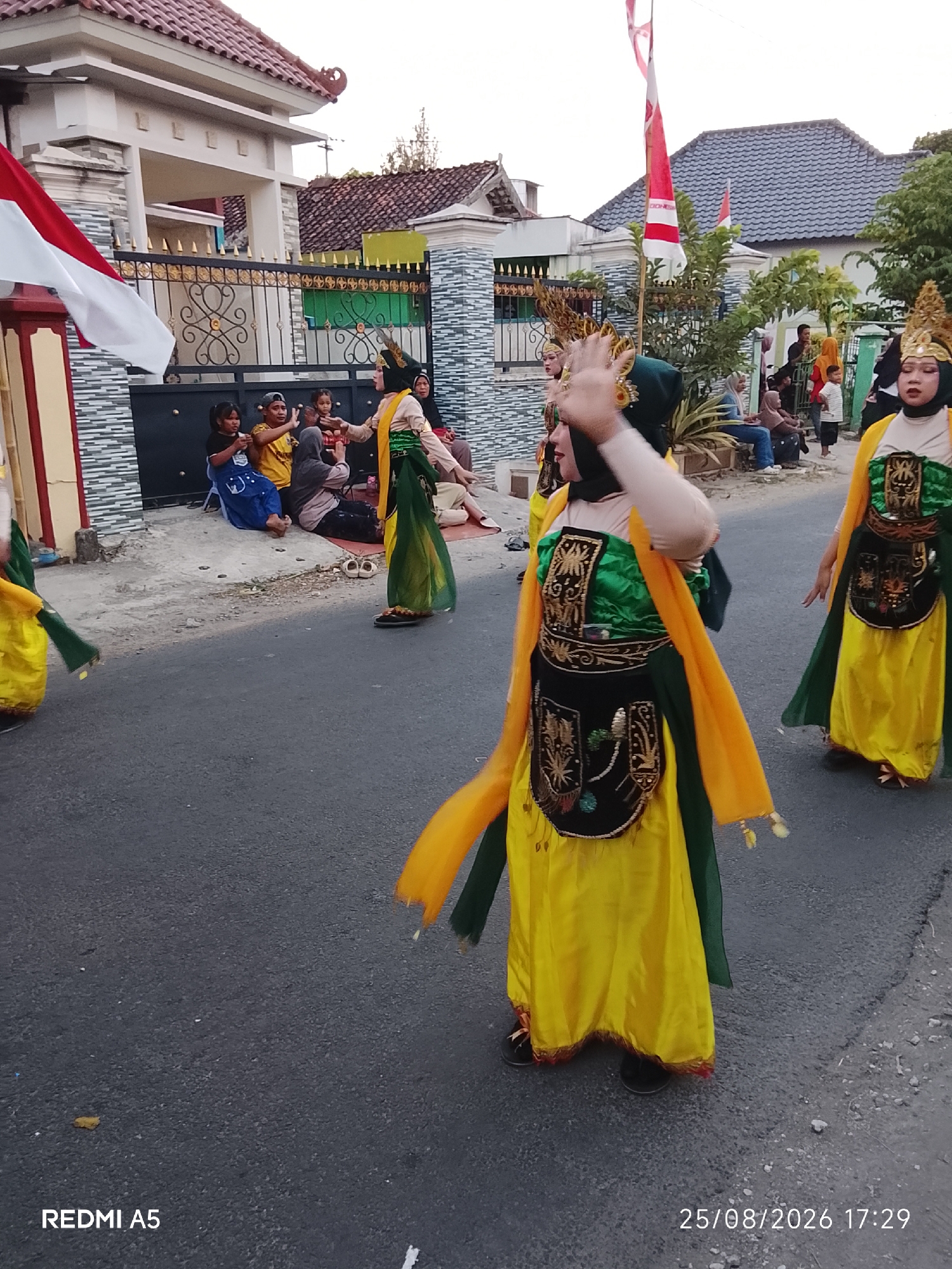 In Semanding, Indonesia, at 17:29 on 08/25/2026, two individuals in vibrant yellow and green traditional dance costumes perform on a paved road. The costumes feature ornate black vests adorned with gold embroidery and beaded fringe, over flowing yellow skirts. One dancer, in the foreground and to the right, is partially obscured by their own yellow sash. The other dancer, to the left, is also in motion, with a yellow sash flowing behind them. Both dancers wear headpieces and appear to be wearing dark shoes. In the background, a group of spectators sits or stands behind a decorative black metal fence. The fence is in front of a building with a tiled roof and white facade, and adjacent to a smaller, darker building. A red and white Indonesian flag flies from a pole on the left side of the frame. The environment appears to be a residential street, with trees and other foliage visible in the distance.