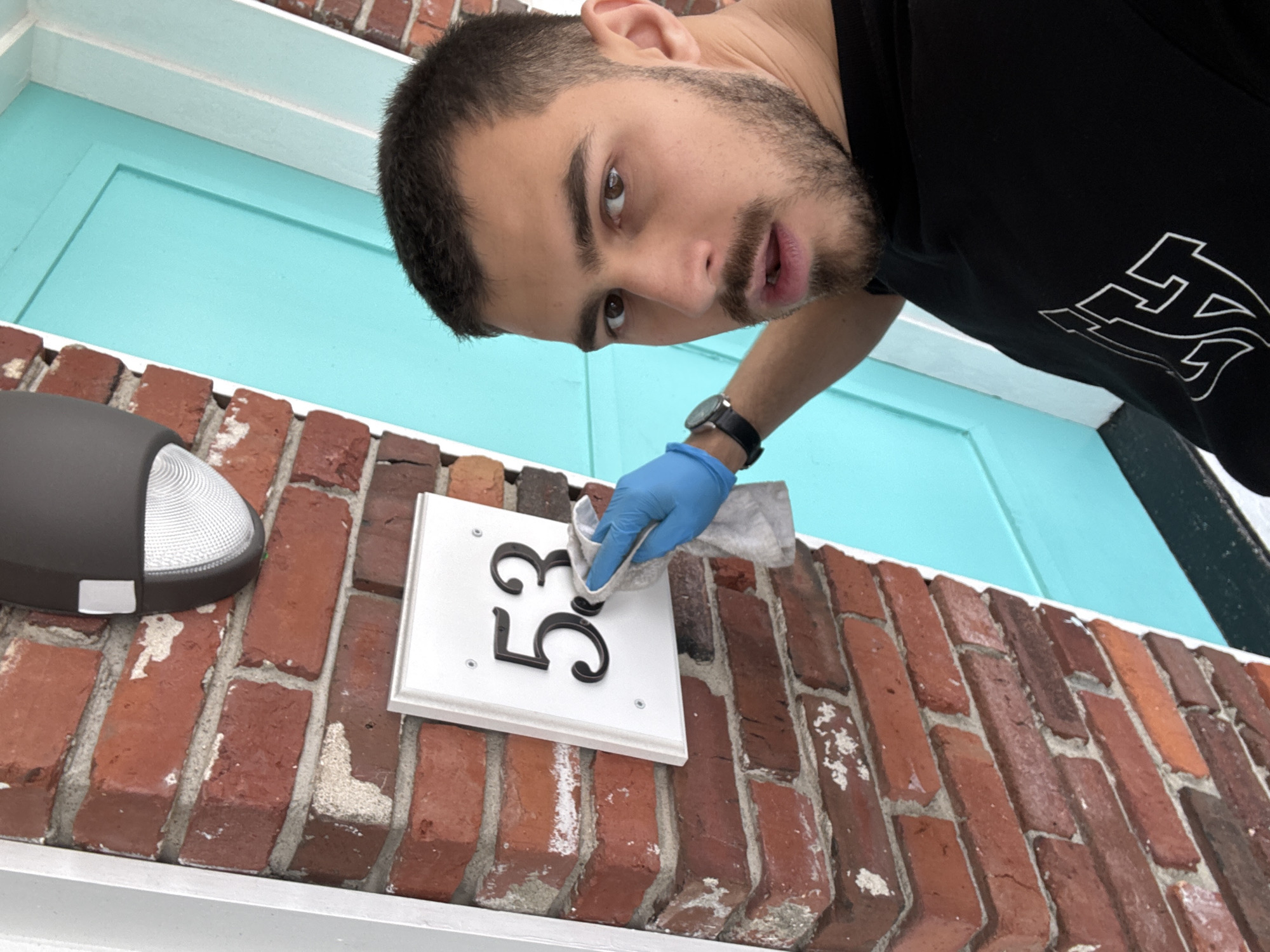 This image depicts a young man engaged in cleaning a house number plaque on the exterior of a building, likely a residence in Hampton, United States of America. The photograph is taken from a low, tilted angle, looking upwards.
The main subject is a man with short dark hair, a beard, and dark eyes, who appears to be in his 20s or 30s. He is wearing a black t-shirt with a white, stylized graphic design on the front. On his left wrist, he wears a dark-strapped watch, and his right hand is covered by a light blue latex or nitrile glove, holding a light-colored cloth or rag. He is looking directly towards the camera with a focused expression.
The activity taking place is the cleaning of a white rectangular house number plaque, which clearly displays the black numbers "53". The man's gloved hand and cloth are positioned over these numbers. The plaque is mounted on a red brick wall, characterized by visible white mortar and some areas of wear or efflorescence. To the left of the plaque, a dark grey outdoor wall light with a textured lens is partially visible. In the background, above the brick wall, a vibrant turquoise-colored door or door frame with white trim provides a contrasting element.
The scene is set outdoors during what appears to be clear daytime conditions, given the bright and even lighting. The visual cues suggest a routine maintenance task.