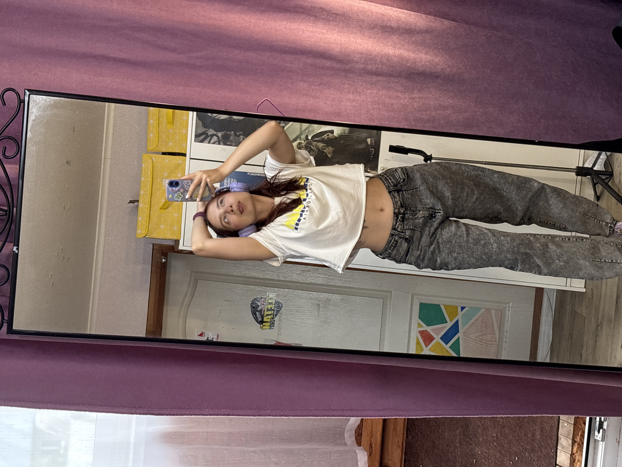 A young woman with shoulder-length brown hair is taking a selfie in a full-length mirror. She is wearing a white crop top with a yellow and blue design on the front and distressed grey jeans. She has on purple headphones and her right hand is behind her head, while her left hand is holding a phone. Her tongue is sticking out slightly. The mirror is framed with black metal scrollwork. Behind her, reflected in the mirror, are two yellow storage boxes with a diamond pattern, a white door with stickers and a geometric design, and a black tripod. The environment appears to be an indoor space, possibly a room.