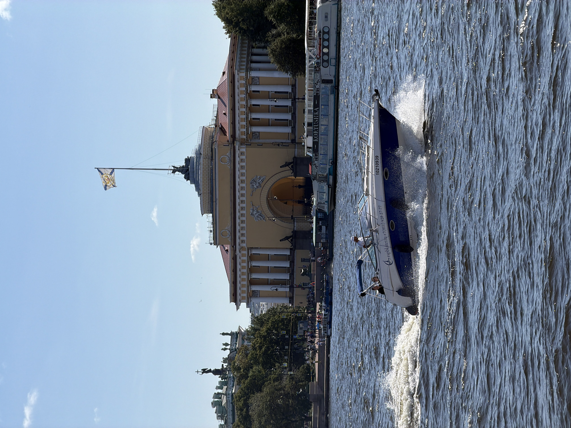 This is a photograph taken on a sunny day in Saint Petersburg, Russia, likely during daylight hours, showing a body of water, possibly the Neva River, with several boats. In the foreground, a white and blue speed boat with a blue canopy is moving rapidly, creating a significant wake. Two people are visible in the boat. Further in the background, a larger tour boat named "KORYUSHKA" is docked or moving slowly. Behind the boats, on the shore, stands a large, light yellow neoclassical building with a red roof, prominent white columns, and an arched entryway. This building is likely the General Staff Building or a similar historical structure on Palace Square, given its architectural style and the presence of a flagpole with a Russian naval or imperial flag flying from its roof. The sky is clear blue with a few wispy clouds. Trees and other buildings can be seen on the left side of the frame, suggesting an urban setting.