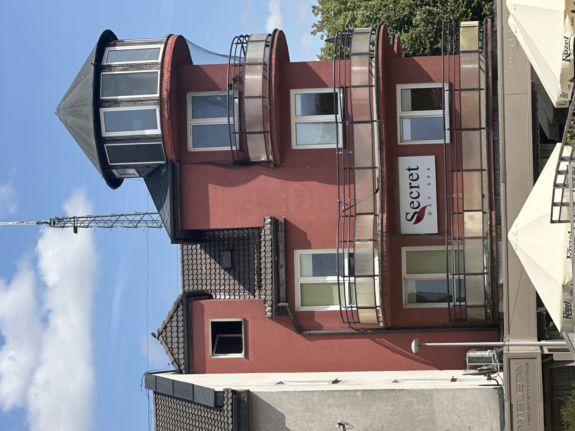 An exterior, eye-level, medium shot captures a red, multi-story building with a distinctive turret-like structure on its upper level. The building is situated in Pruszcz Gdański, Poland, and appears to house a "Secret Day Spa," indicated by a prominent sign on the facade. Below the spa, a sign for "Kameleon restaurant & drinks" is visible. The architecture features multiple balconies with glass and metal railings. A tall communication tower stands behind and to the left of the building. The sky above is blue with scattered white clouds, and a lush green tree is visible to the right, suggesting a daytime setting. A bird is perched on the rooftop.