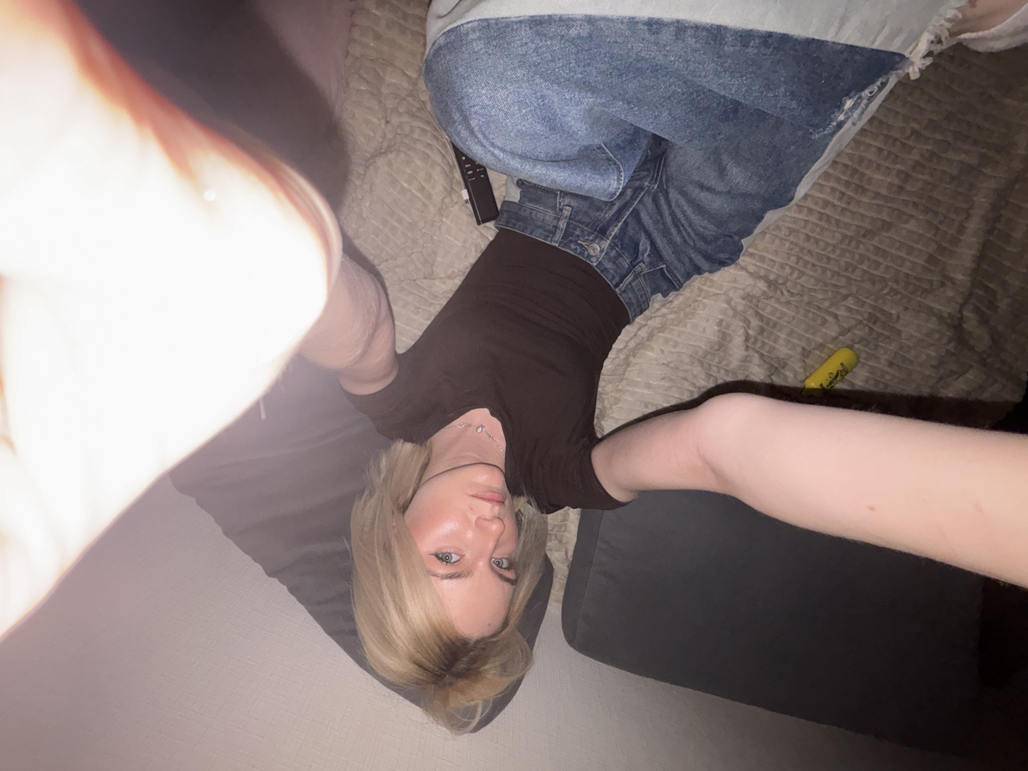 A woman with blonde hair is lying on a bed, wearing a brown shirt and jeans. A TV remote and a yellow object are on the bed beside her. She is taking a selfie from an upside-down perspective, and the photo is overexposed on the left side. The location is indoors, likely in a private residence. There is no indication of the location being in Vsevolozhsk, Russia.