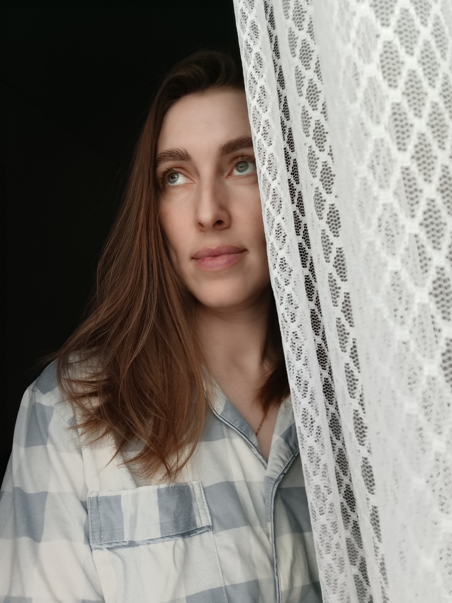 A woman with long brown hair is partially obscured by a white lace curtain. She is wearing a blue and white plaid shirt. The background is a dark, indeterminate space. The location is Chaikovsky, Russia.