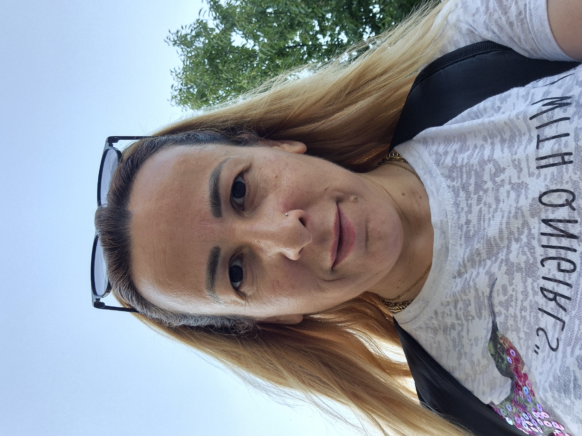 A medium shot, selfie-style, portrait of a woman with blonde hair and sunglasses perched on her head is captured outdoors against a backdrop of a clear sky and a green tree. The woman is looking directly at the camera, her head slightly tilted. She has a gold necklace visible around her neck and is wearing a white t-shirt with a print of a hummingbird and sequins, as well as a black backpack. The image appears to have been taken during the day, with natural lighting. The location is described as Unknown, Unknown, implying no specific geographical information is available.