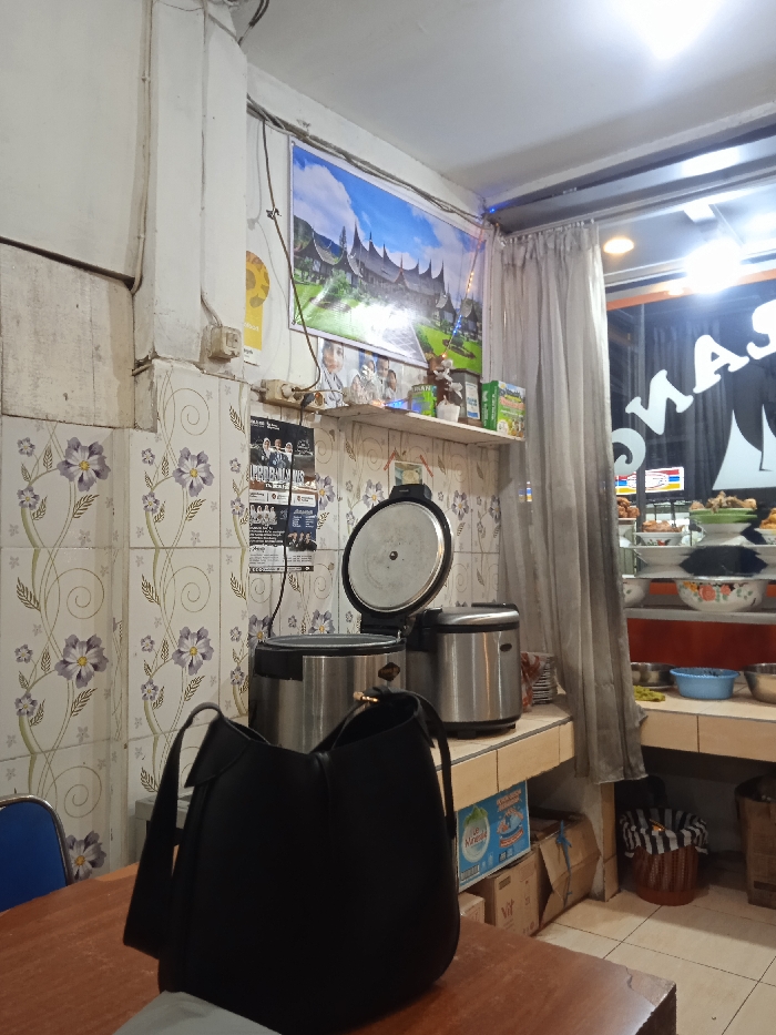 The scene is an interior shot of a restaurant or shop in Kecamatan Kartasura, Indonesia. The image appears to be taken near a counter, and there is a white tiled wall covered in a floral pattern to the left, and a window to the right. There is a black purse on the counter, in front of an open rice cooker and an electric rice cooker. On the wall to the left of the rice cookers there is a poster. To the right of the rice cookers there is a window looking out onto a street, which has a red and white sign with the word “MAS” on it. The window is in a room with a white tiled wall and a counter made of light-colored wood. The window is partially obscured by a white curtain, with brown and blue decorations above it. The room has a white ceiling with a single light fixture.
