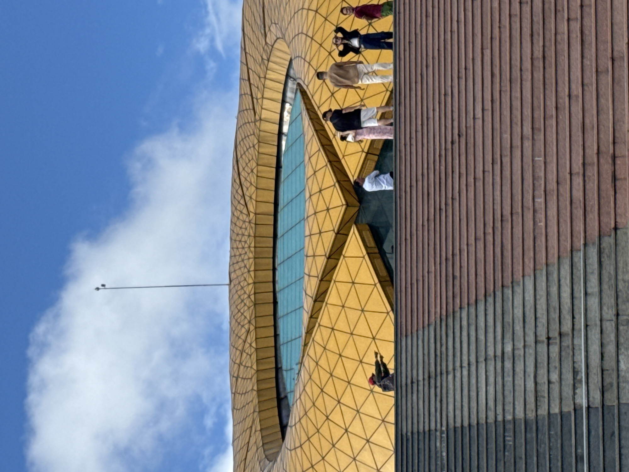 The image depicts the exterior of the Da Lat Flower Garden (Vườn Hoa Đà Lạt) in Da Lat, Vietnam. A group of people are visible standing on a set of wide, multi-colored stairs that lead up to a large, modern building with a distinctive golden, honeycomb-like roof structure. The building features a prominent blue glass-covered area in its center, resembling an eye.
In the foreground, the stairs are a prominent feature, with sections appearing in shades of gray and reddish-brown. Several individuals are scattered across the upper portion of these stairs. A lone figure wearing a red cap and dark clothing is positioned on the left, seemingly pointing outwards. To the right of this figure, a group of six individuals are more closely clustered, observing the building or the surroundings. One woman in the group is wearing a pink floral dress, and a man next to her is in a black shirt and white shorts. Another man in a tan shirt and white pants stands to the right of them. A woman in a black jacket and dark pants is also present, with her hands near her head. A man in a red shirt and green pants is visible on the far right of this group.
The background is dominated by the striking golden roof of the Da Lat Flower Garden building, which has a futuristic and organic design. A tall pole with some equipment at the top extends into a bright blue sky with scattered white clouds. The overall scene suggests a sunny day, with the architecture of the building and the presence of visitors indicating it is a public attraction.