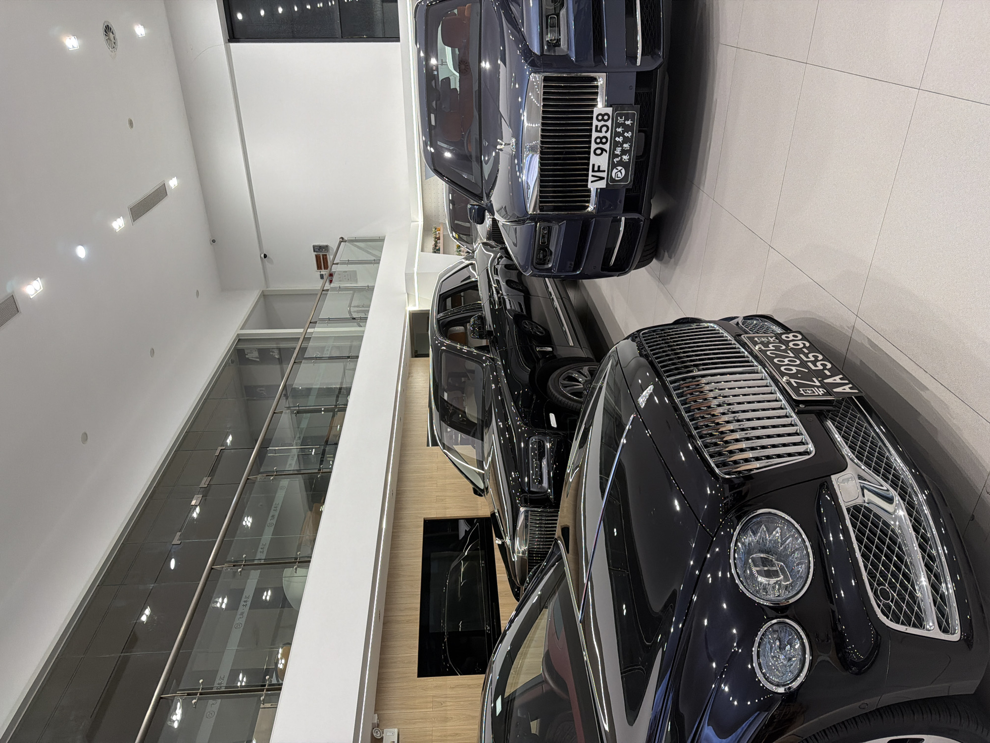 The image depicts the interior of a luxury car dealership, likely a Rolls-Royce or Bentley showroom, located in Dongguan, China. In the foreground, the front of a black Bentley is visible, featuring its distinctive grille and headlights. To the right, a dark blue Rolls-Royce Cullinan is parked, with its license plate reading "VF 9858" and Chinese characters below. Behind the Cullinan, another Rolls-Royce, possibly a Phantom, is partially visible, with a black Rolls-Royce Cullinan parked further back on the left side of the image. The showroom has a clean, modern aesthetic with polished tiled floors and large windows reflecting the ceiling lights and the upper levels of the building. A glass railing separates the showroom floor from an upper walkway, where more of the building's interior architecture, including light fixtures and ventilation systems, can be seen. The overall impression is one of opulence and high-end automotive display.