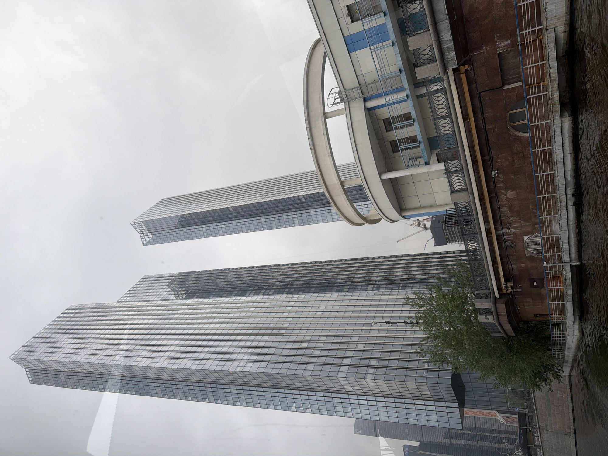 The image shows a view of the Moscow International Business Center (Moscow City) in Moscow, Russia. The buildings are tall skyscrapers with glass facades, reflecting the cloudy sky. The structures include the Evolution Tower with its distinctive spiral shape, and other modern office towers. A section of a waterfront or embankment is visible along the right edge of the image, with part of a river visible, and buildings above it, suggesting the view is from a low angle, possibly from a boat on the river.