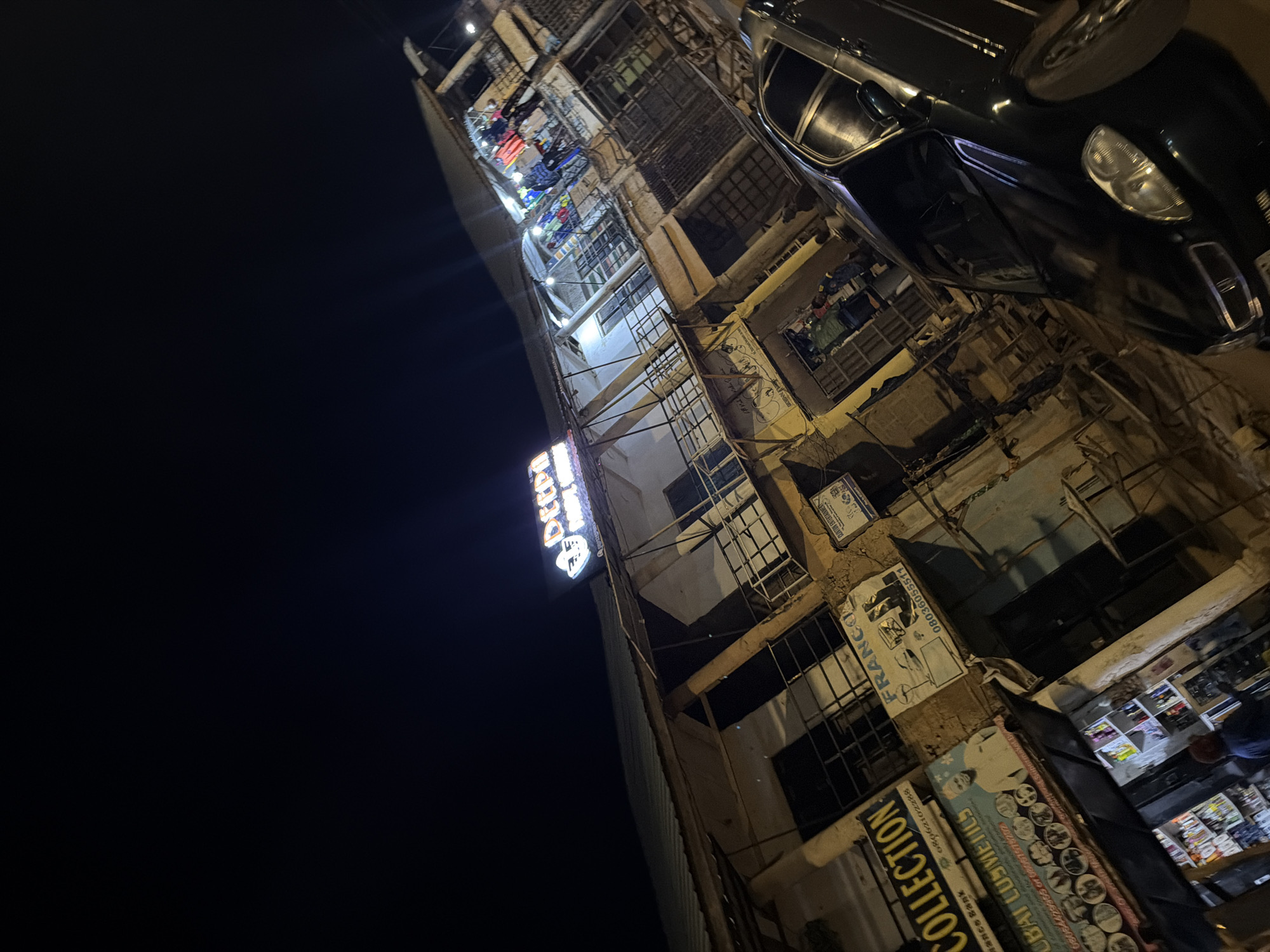 The image captures a nighttime scene in Bukuru, Nigeria, focusing on a multi-story building housing various shops. The building has a slightly weathered facade, with balconies and grilled windows. Several signs are visible, including one for "DEEPM," a luminous sign at the top of the building, another for "COLLECTION," and a third for "DUBAI COSMETICS." One sign reads "FRANCO" with a phone number. Inside some of the shops, shelves stocked with products are illuminated. A dark-colored sedan is parked in front of the building, partially obscuring the lower level. The street is dimly lit, typical of a nighttime urban environment. The image is taken at an angle, giving a dynamic perspective of the scene.