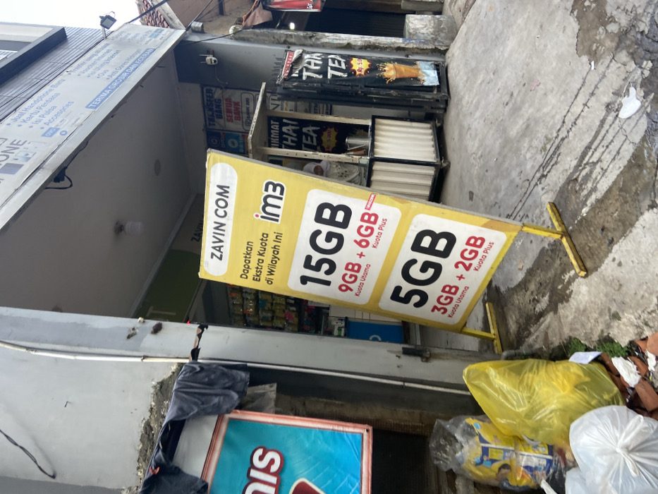 A yellow sign with the words "Zavin Com" and "im3" on it is leaning against a building. It is advertising data plans for a mobile phone provider. The sign lists data plans for 15 GB, 9 GB, and 5 GB. There are a few garbage bags in the foreground, and a partially obscured sign in the background. The location is in the city of Kecamatan Cimahi Selatan, Indonesia at latitude -6.932 and longitude 107.564. The ground is concrete and the sign is standing on a metal frame. A person is partially visible in the background, standing in front of a counter.