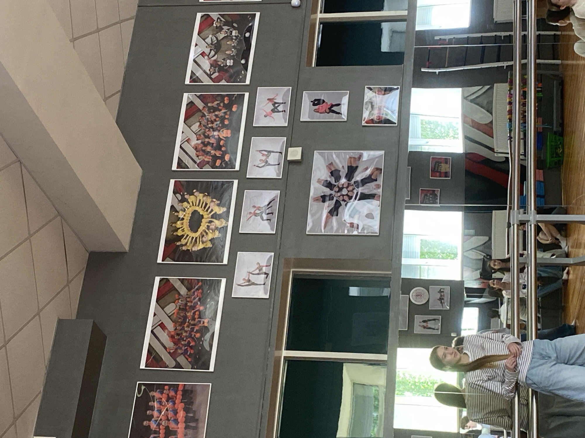 The image depicts the interior of a dance or gymnastics studio in Votkinsk, Russia, characterized by its large mirrors, wooden floor, and barre equipment. The walls are adorned with numerous framed photographs, primarily showcasing groups of young people in athletic poses, suggesting a focus on dance or performance. Prominently displayed are large photos of groups in coordinated outfits, some forming circular patterns. Smaller framed images of individuals and pairs in dynamic dance positions are also visible. In the foreground, a young woman with long brown hair, wearing a striped long-sleeved shirt and light blue wide-leg pants, stands near a barre, looking towards the left of the frame. Several other individuals, mostly young women, are seated or standing in the background, some reflected in the mirrors. The studio appears well-lit, with windows visible in the reflections, suggesting natural light supplemented by artificial lighting. The overall impression is of a training or performance space dedicated to movement arts.