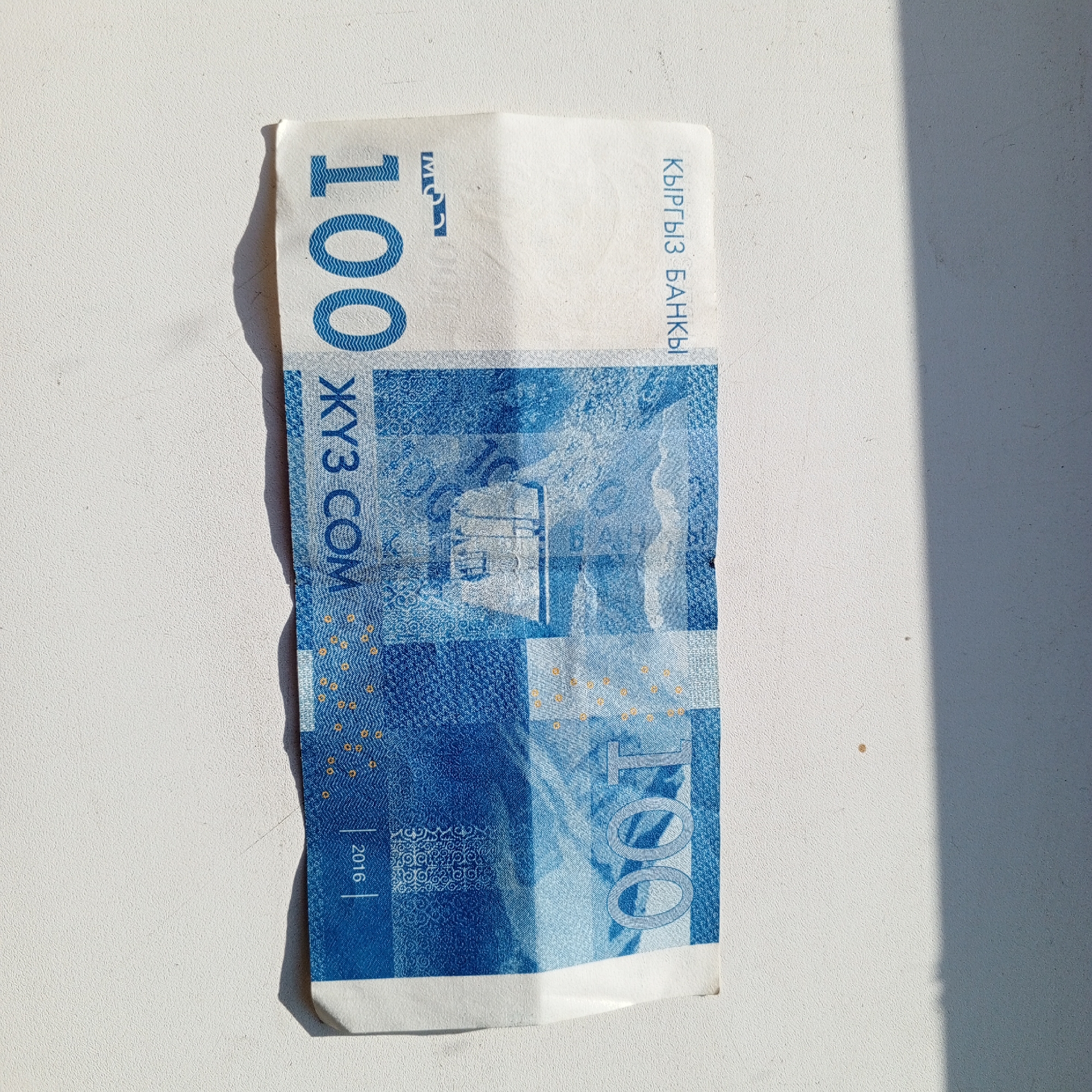 The image shows a single banknote, folded vertically, lying on a textured white surface. The banknote has a predominantly blue and white design with some yellow elements. The text on the banknote is in Cyrillic and appears to be in a foreign language. The numeral "100" is prominently displayed in blue. The image is captured in daylight, with a shadow cast across the right side of the scene, suggesting an outdoor or window-lit environment. The banknote is identifiable as a Kyrgyzstani Som. No individuals or specific city landmarks are visible. The location in Sayanogorsk, Russia, is not indicated by any visual cues within the image itself.