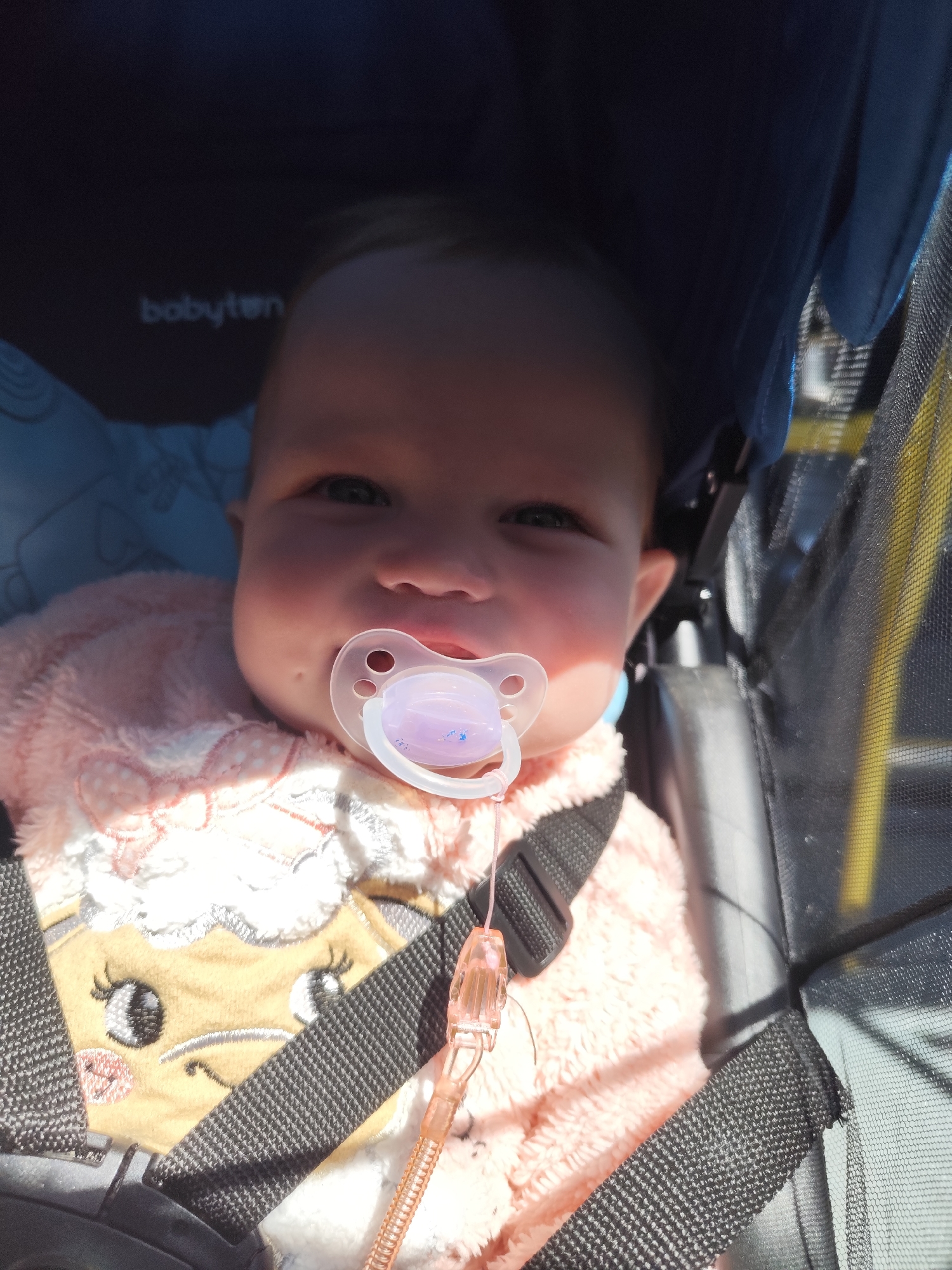 An infant is pictured sitting in a dark-colored stroller under bright sunlight. The infant has fair skin, dark eyes, and a soft facial expression, looking directly forward. A clear plastic pacifier with a pink/purple silicone nipple is in the infant's mouth, connected by a pink ridged clip to the garment. The infant wears a plush, pink and white garment featuring an embroidered character with a beige face, black eyes, and white textured "wool" on the torso.
The infant is secured in the stroller by black webbing harness straps. The stroller's dark blue fabric canopy is visible in the upper left, displaying a white "babyten" logo. A patterned, light blue lining is seen on the stroller's interior to the infant's left. The right side of the stroller features black mesh fabric, through which a yellow vertical element is partially visible. Shadows cast by the stroller structure and harness indicate direct, strong overhead sunlight. The image does not contain any discernible visual information to identify the location as the city of Chaikovsky, Russia, or any other specific geographical area.