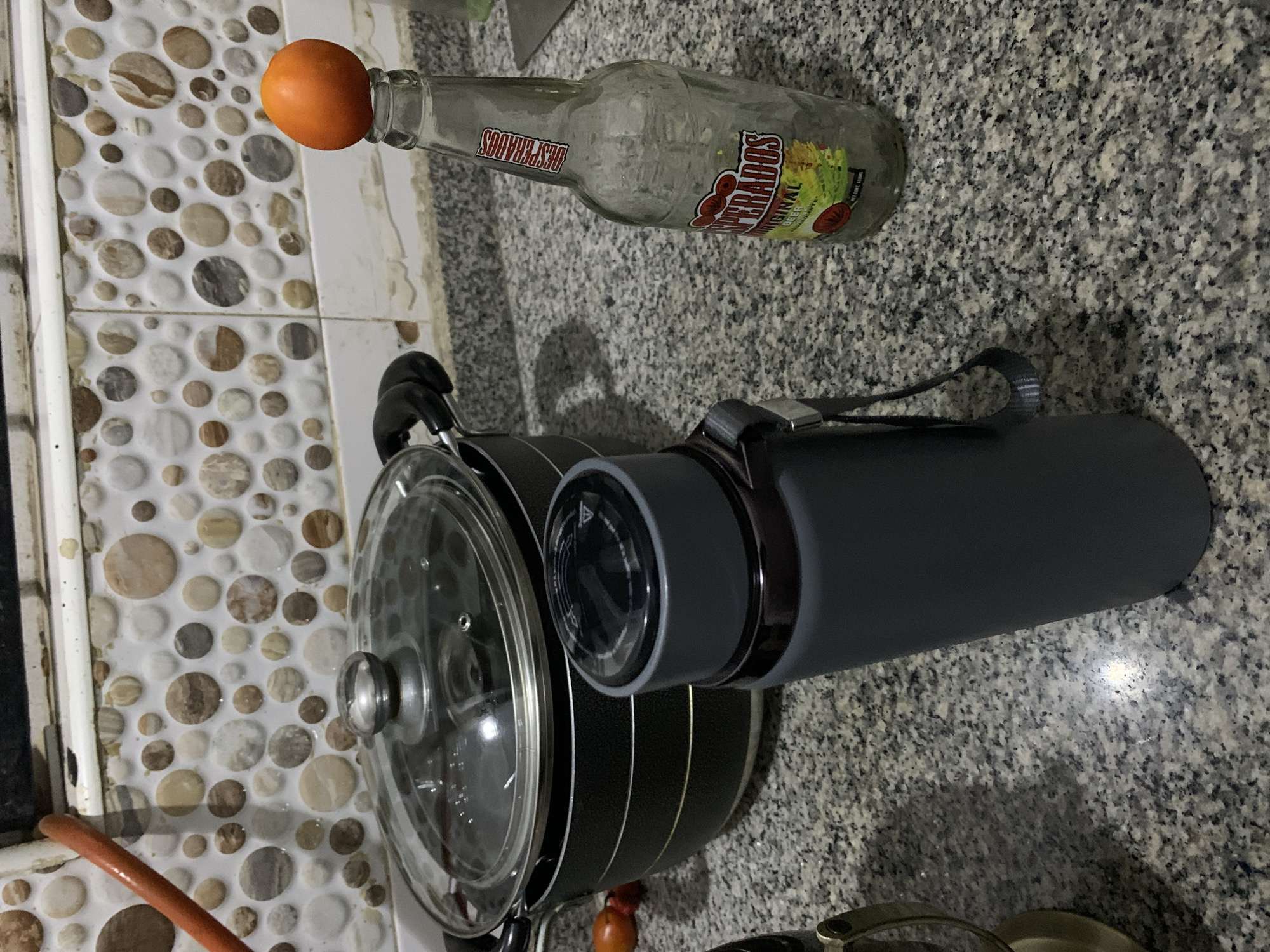 This image captures a still life arrangement on a kitchen counter in Gwarinpa, Nigeria. The scene is dominated by a grey speckled granite countertop and a white tiled wall featuring a decorative mosaic pattern of various-sized, round, smooth stones in shades of brown, grey, and cream.
In the upper portion of the image, an empty glass bottle of "DESPERADOS ORIGINAL BEER" rests on its side on the countertop, with a small orange precariously balanced on its neck. The bottle's label is clearly visible, showing the brand name and the words "ORIGINAL BEER".
Towards the foreground and center, a black cooking pot with a clear glass lid is visible, accompanied by a dark grey thermos-style bottle with a black cap and a visible dial. The dial features some unreadable symbols and a small triangle icon. A black strap is attached to the thermos. Parts of other kitchen items, including handles, are also faintly visible around the pot.
There are no people present in the image, and no specific activity is taking place. The lighting suggests an indoor setting, likely artificial light. The overall impression is a casual, everyday kitchen scene.