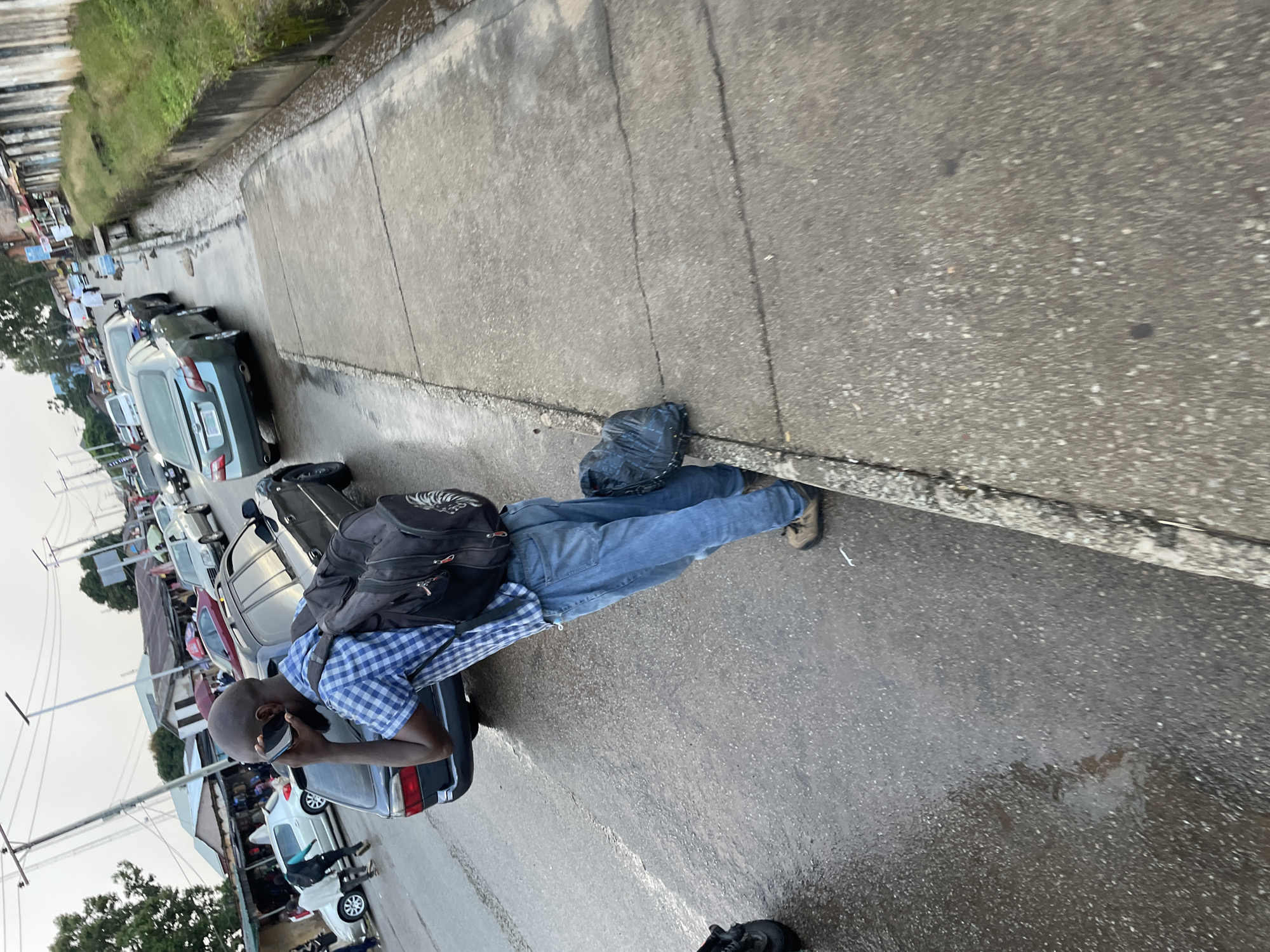 A bald Black man wearing a blue and white checkered short-sleeved shirt, blue jeans, and brown shoes stands on the side of a wet road, talking on his phone. He has a black backpack slung over his right shoulder. A black trash bag is on the ground near his feet. The road is busy with traffic, including a gray sedan and other vehicles. Buildings line the right side of the road, with some appearing to be shops or market stalls. Overcast sky. The scene appears to be in Jos, Nigeria.