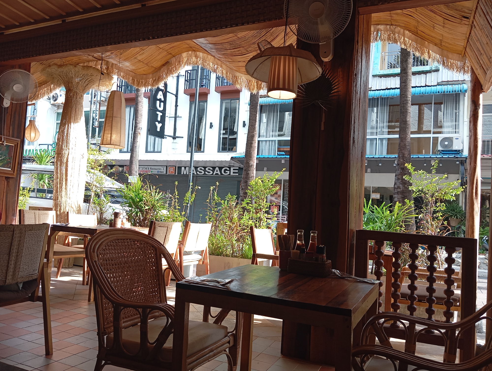 The image depicts an outdoor seating area of a restaurant in Karon, Thailand. The foreground features a wooden table set with condiments in a basket, a napkin, and cutlery. Two wicker chairs with light-colored cushions are positioned at the table. In the background, several other tables and chairs are visible, some with woven backs. Lush green plants are strategically placed throughout the seating area, adding to the tropical ambiance. Through an opening, a street view of Karon is visible, with buildings, palm trees, and people walking by. The architecture and decor suggest a relaxed, open-air dining experience, characteristic of a beachside resort town. The overall scene is bathed in natural light, indicating it was taken during the daytime.