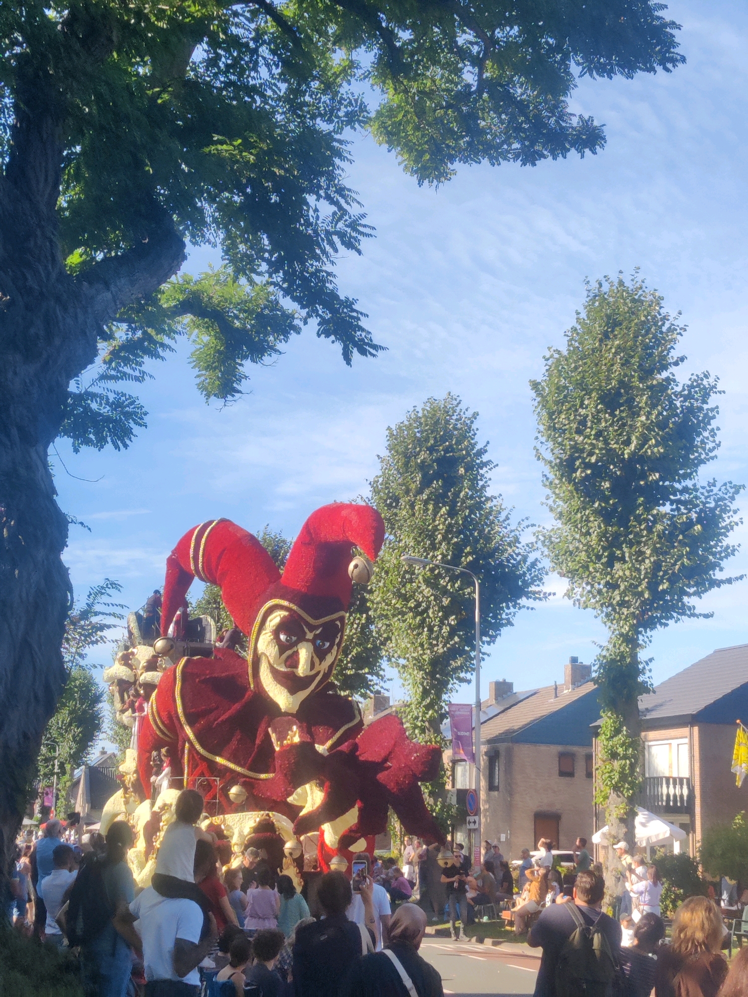 This is a daytime, eye-level, medium shot of a parade in Zundert, Netherlands, likely during the Bloemencorso. The main subject is a large float shaped like a menacing jester with a red and gold costume, exaggerated facial features, and a tall, pointed hat. The float is adorned with what appears to be flowers and other decorative materials. It is being pulled by a vehicle, partially visible behind the jester.
A crowd of spectators lines the street, with people of various ages watching the parade. Some individuals are standing, others are sitting on the curb or on portable chairs. Many are holding up mobile phones to capture images or videos of the event. A child is being carried on someone's shoulders for a better view.
The environment consists of a tree-lined street with houses and buildings in the background. Tall, leafy trees frame the top and sides of the image. The sky is blue with wispy white clouds. The overall atmosphere appears festive and communal, with people gathering to celebrate the parade. The presence of a street lamp and traffic signs suggests an urban or suburban setting.