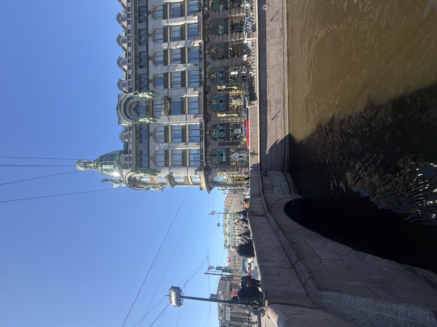 The image presents a vibrant daytime scene in Saint Petersburg, Russia, centered on the iconic Singer House (Dom Knigi) and a canal. The impressive multi-story building occupies the left side of the frame, showcasing ornate Art Nouveau architecture. Its light-colored facade is punctuated by numerous windows, some arched, and adorned with intricate sculptural details and balconies. A prominent feature is the building's distinctive green glass and metal dome, topped with a large sculptural winged figure. Additional green-patinated statues and decorative elements are visible across the building's exterior.
To the right of the building, a wide canal or river flows with brownish-green water, reflecting the clear blue sky and shimmering with sunlight. A sharp, dark shadow extends across a significant portion of the water, indicating bright, direct sunlight. A robust stone embankment, constructed from large, rough-hewn blocks, separates the building's street from the canal. This embankment features a pronounced curve, leading into what appears to be an archway or underpass.
Several people are visible along the embankment, some walking and others standing, enjoying the sunny weather. Overhead, utility wires crisscross the expanse of the clear, azure sky. In the lower right foreground, a street pole is visible, bearing a white rectangular sign with the Cyrillic word "СТОЛ" (meaning "table" or referring to a dining establishment/public utility marker) printed on it. The scene conveys a bright, clear day, likely late morning or early afternoon, with a bustling urban atmosphere.