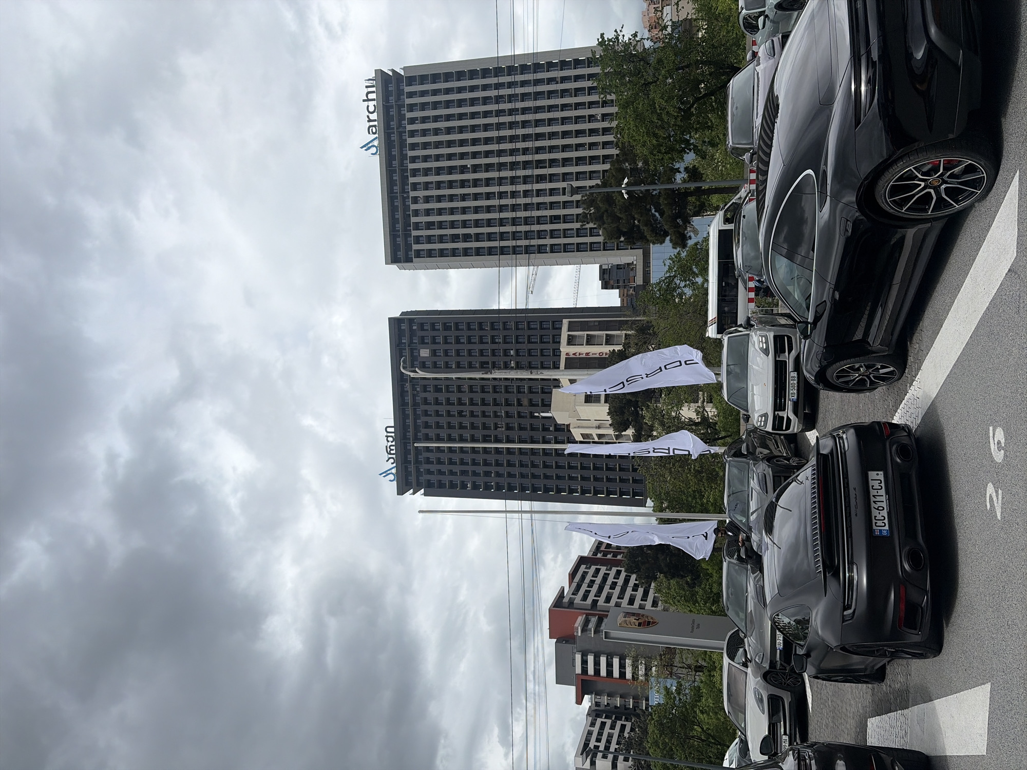 The image captures a scene in Tbilisi, Georgia, likely near a Porsche dealership, showcasing the brand's vehicles against a backdrop of modern architecture. The sky is overcast, suggesting a cloudy day. Two tall buildings dominate the background, one featuring the "archi" logo at its top. Several white flags bearing the "PORSCHE" logo are prominently displayed near the foreground. A row of Porsche cars is parked along the street, with one particularly visible car having the license plate "CC-611-CJ." Other vehicles in the vicinity show registration plates such as "XB-580-BB-" and "-713 09-MA". A Porsche dealership sign is visible, reading "Porsche Centre Tbilisi". The image composition is slightly tilted.