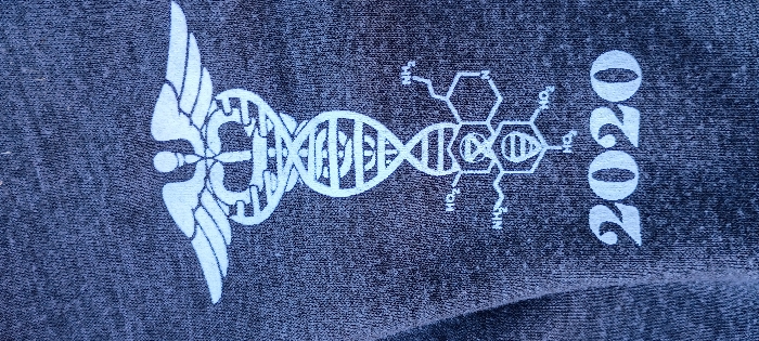 This close-up image, documented in Rohnert Park, United States, displays a dark grey heathered textile, consistent with a garment material. A white graphic design is screen-printed vertically down the center of the fabric. The design features a Caduceus symbol at the top, followed by a double helix structure representing DNA. Below the DNA, a complex chemical molecular diagram is depicted, including visible 'NH2' and 'HO2' functional groups. At the base of the graphic, the numerals '2020' are printed. The textile exhibits minor creases, indicating it is not perfectly flat, and the background beyond the fabric itself is entirely obscured. No individuals or external environmental details are discernible within the frame.