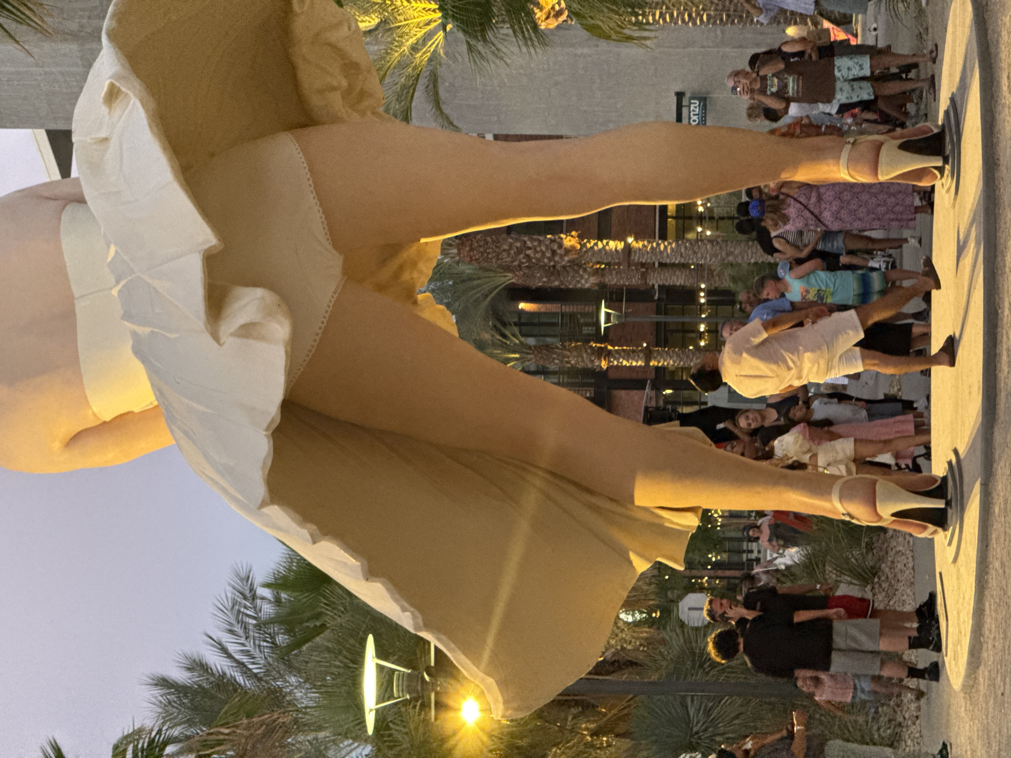 The image captures a low-angle, close-up view of a large sculpture of Marilyn Monroe's legs and skirt from the iconic "Seven Year Itch" pose. The sculpture stands in an outdoor public space in Palm Springs, California, identifiable by the palm trees and arid landscape. Several people are gathered around the base of the sculpture, observing it.
The sculpture itself features Monroe's legs clad in white heels, with her dress billowing upwards. The lighting suggests it is either early morning or late evening, with warm artificial lights illuminating the scene. The background shows a modern building with a sign that appears to say "onzu." The overall atmosphere is that of a tourist attraction or public art installation.