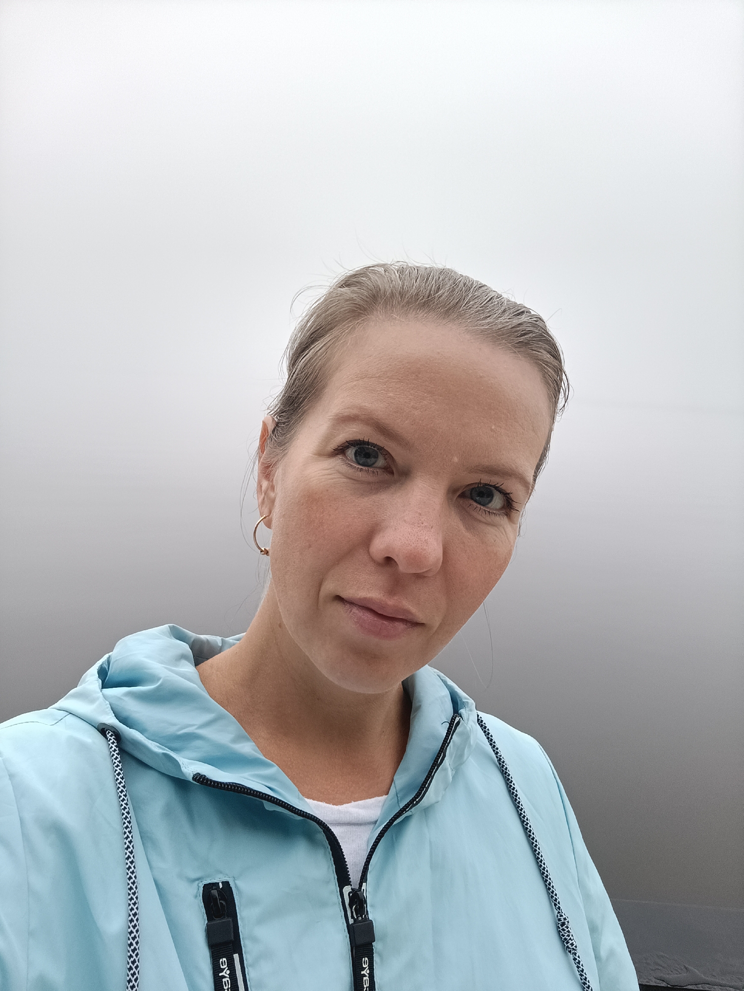 A headshot-style portrait captures a woman looking directly at the camera. She has fair skin with some freckles, light-colored hair pulled back, and striking blue eyes. She wears small gold hoop earrings. Her expression is neutral and direct.
She is dressed in a light blue hooded jacket, typical of a windbreaker, with a black zipper and black and white braided drawstrings around the hood. A white t-shirt is visible underneath. On the zipper pull tabs, the white text "SYEAG®" is visible on black straps.
The background is an expansive, undifferentiated field of light gray, fading to white at the top, strongly suggesting very foggy or heavily overcast conditions. No horizon or specific landscape features are discernible, indicating an open outdoor environment, potentially a large body of water or open terrain. The muted, even lighting points to daytime. The image appears to be a posed selfie or close-up portrait. The location context of Chaikovsky, Russia, aligns with a potential setting near the Kama River under foggy weather.