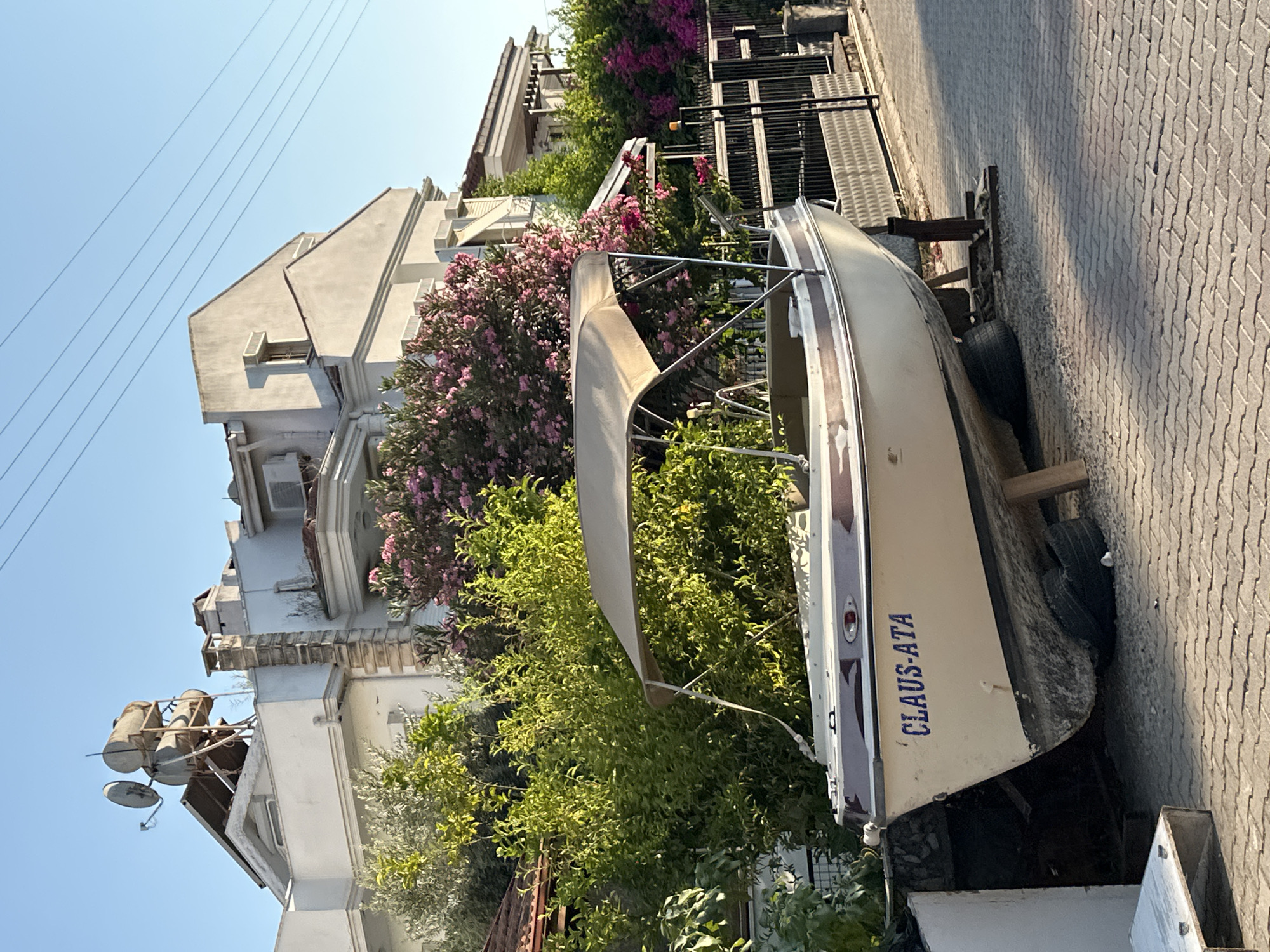 A boat is parked on a paved area next to buildings, possibly in a coastal town. The boat, labeled "CLAUS-ATA," is resting on a trailer with visible tires. Behind the boat, there are lush green trees and blooming pink flowers, suggesting a warm climate and a well-maintained environment. The buildings in the background appear to be multi-story structures with balconies and architectural details characteristic of Mediterranean or Middle Eastern regions. One building has what looks like satellite dishes and water heaters on its roof. The overall scene is bathed in bright sunlight, indicating it is daytime, likely late morning or afternoon. The paving stones suggest an outdoor setting, possibly a street or courtyard. The presence of the boat on a trailer implies it might be near a body of water, ready for launch, or being stored. The visual information, combined with the prompt's location context of Fethiye, Turkey, paints a picture of a Mediterranean coastal town on a sunny day.