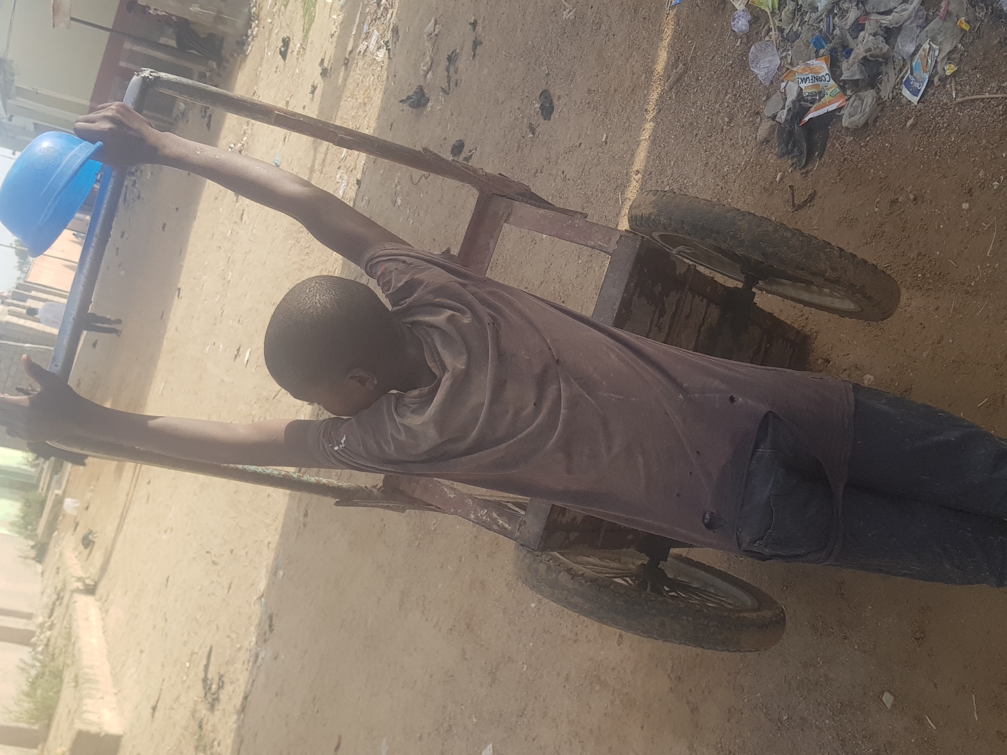 A young person is pulling a metal cart with two wheels on a dirt road in Dabawa, Nigeria. The individual is wearing a brown shirt and dark pants, with arms outstretched to hold the cart handles. On the cart is a blue plastic bowl. The ground is covered in dirt and scattered debris, including plastic bags and paper. Buildings are visible in the background.