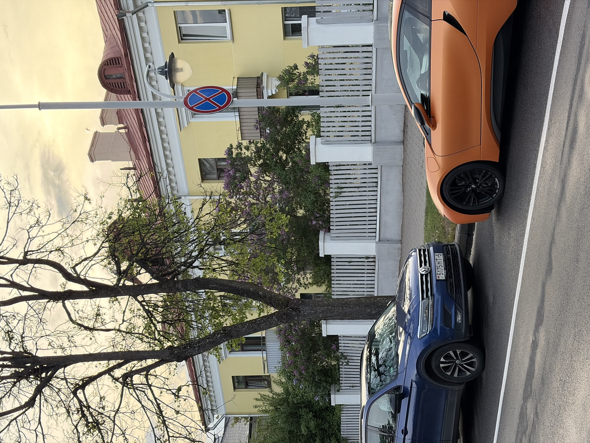 The image depicts a street scene in an urban area. A blue Volkswagen and an orange BMW are parked along the curb. A street sign indicating "No Stopping" is visible. A tree with sparse foliage stands near the vehicles. The surrounding architecture includes a yellow building with a red roof, and a white picket fence. The environment appears to be a residential area during daylight hours.
