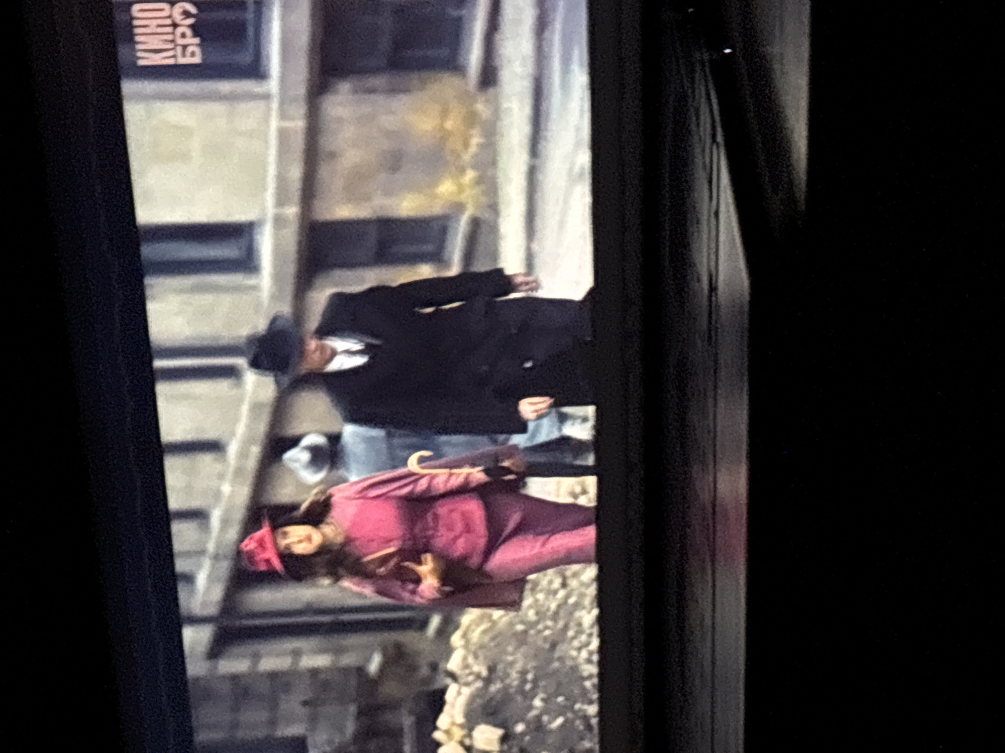 The scene shows a movie being filmed in what appears to be a city setting. A woman in a bright pink suit and matching hat is walking down a street, holding an umbrella. A man in a dark coat and hat is walking slightly ahead of her. Another man in a lighter-colored hat and jacket is standing behind them. The backdrop includes buildings with windows and a sign with the word "КИНО". The location is implied to be Budva, Montenegro, based on the "Kino Budva" logo at the top left. The street appears to be paved, and there are some stones on the side.